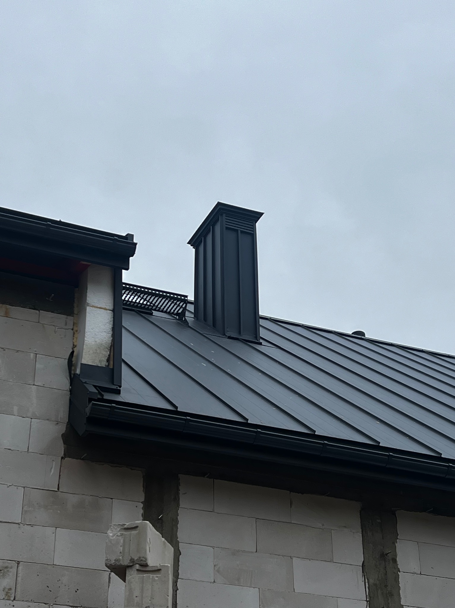 Szary dach z blachy trapezowej, czarny komin i orynnowanie na tle pochmurnego nieba, widoczne bloczki betonowe ściany budynku w trakcie budowy.