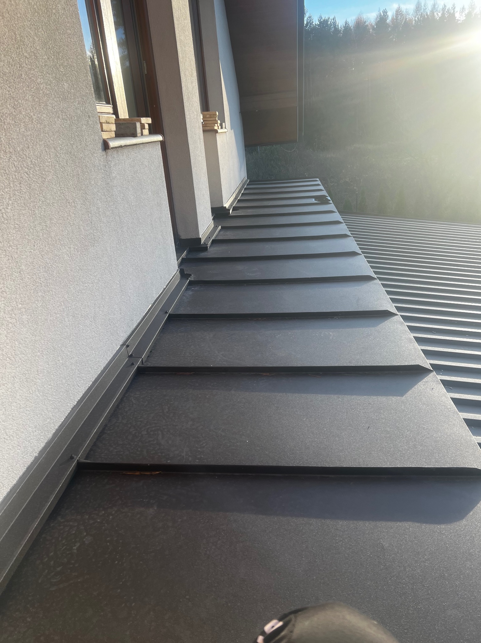 Nowoczesny, grafitowy dach panelowy z widocznymi mocowaniami, przylegający do jasnej ściany budynku z oknami, w tle zalesione wzgórza w porannym słońcu.