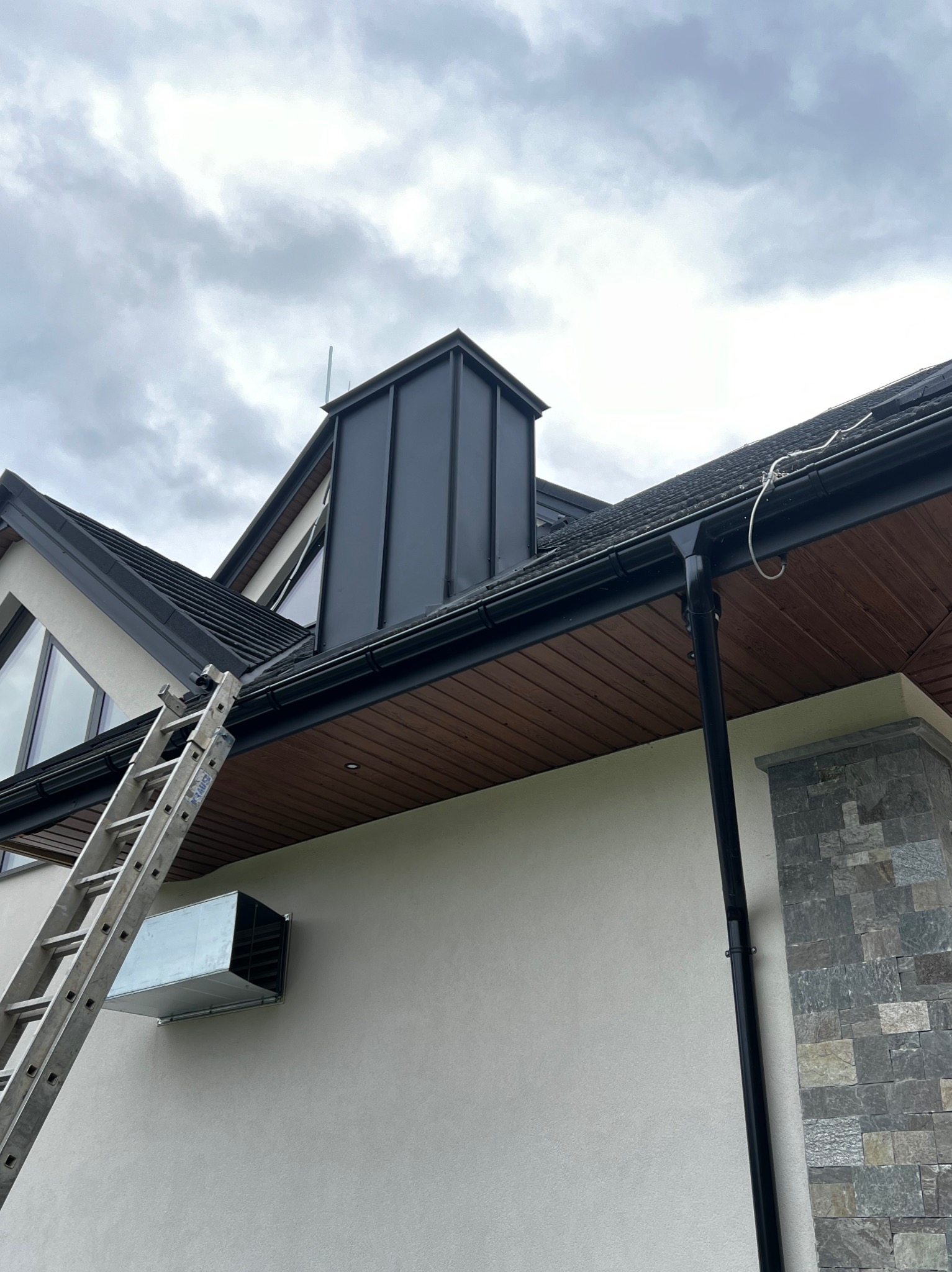 Dom z ciemnym dachem, czarną rynną i kominem z blachy, drabina oparta o ścianę, pochmurne niebo w tle.