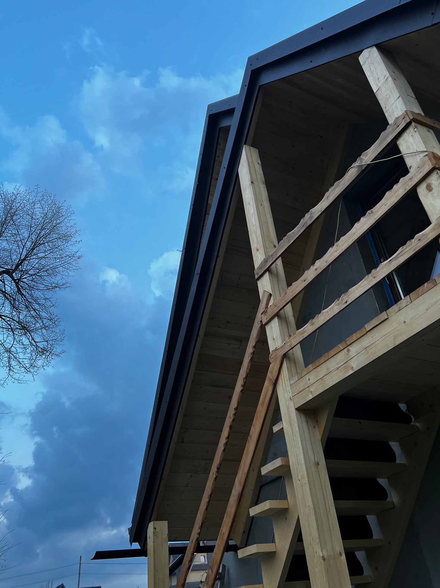Widok z dołu na fragment drewnianej konstrukcji dachu z czarnym orynnowaniem, częściowo osłoniętej drewnianym rusztowaniem, na tle błękitnego, pochmurnego nieba.