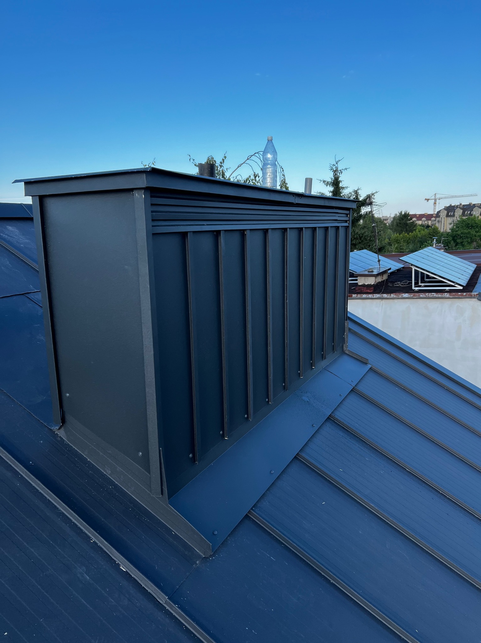 Szary komin wentylacyjny na nowym, ciemnoniebieskim dachu z blachy na rąbek stojący, na tle błękitnego nieba i zabudowań miejskich z żurawiem budowlanym.