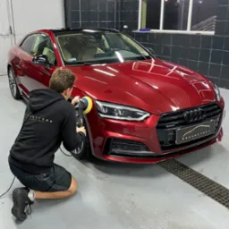 Pracownik w czarnej bluzie z kapturem poleruje czerwoną karoserię Audi A5 za pomocą polerki rotacyjnej Rupes, widoczne logo Krashevsky Auto Spa na grillu samochodu, tło stanowią grafitowe płytki...