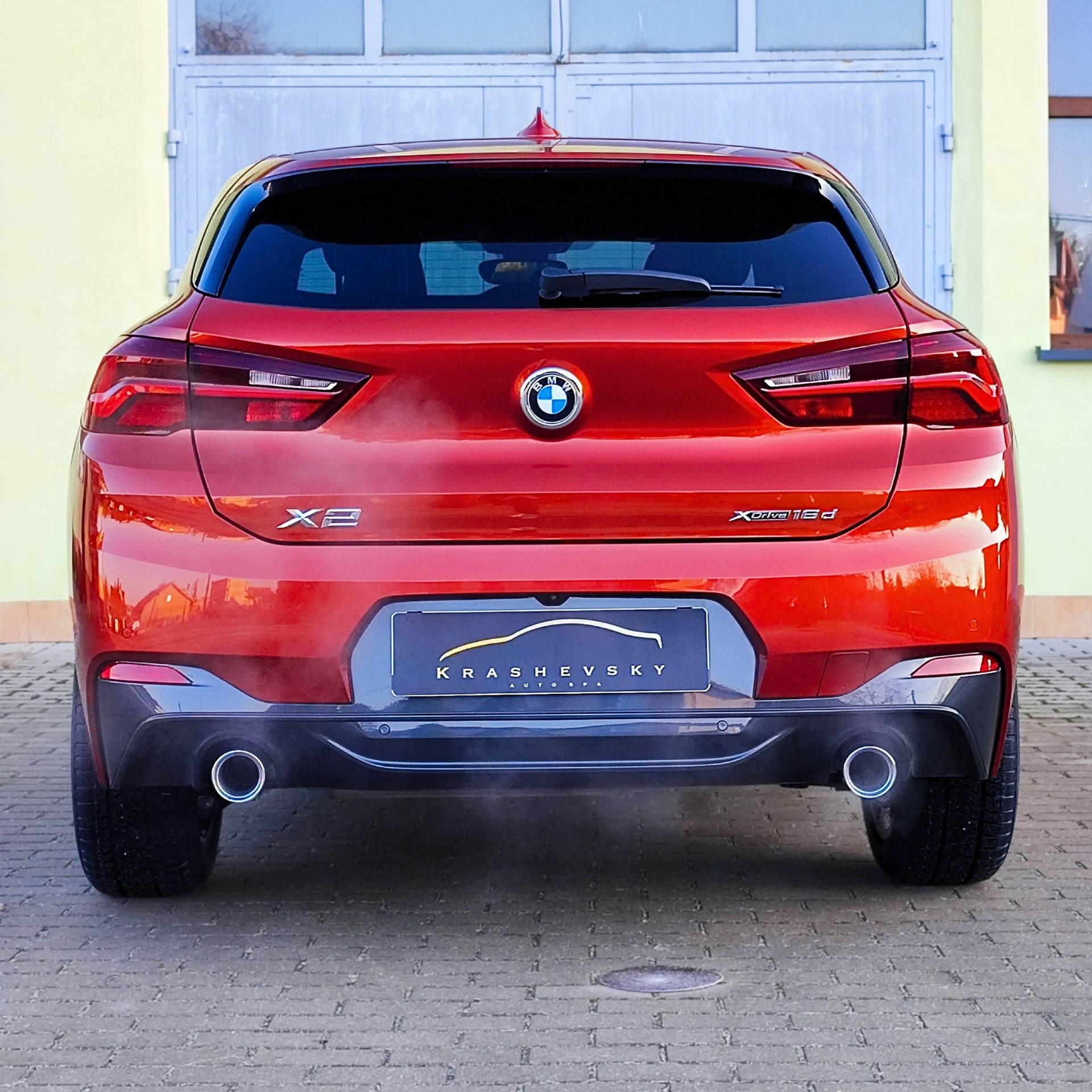 Czerwony samochód BMW X2 xDrive18d, widok z tyłu, z widocznym logo Krashevsky Auto Spa na tablicy rejestracyjnej, stojący na kostce brukowej przed garażem.