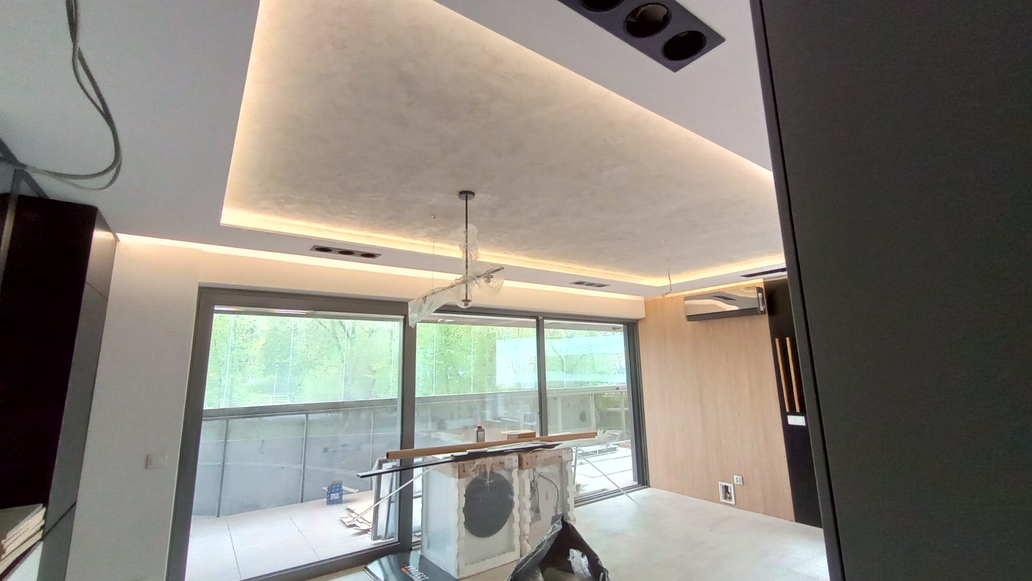 Wnętrze w trakcie remontu z widocznymi przygotowaniami do prac wykończeniowych, zabezpieczona folią lampa sufitowa, sufit z oświetleniem LED i widoczną strukturą, duże okna przesuwne z widokiem...