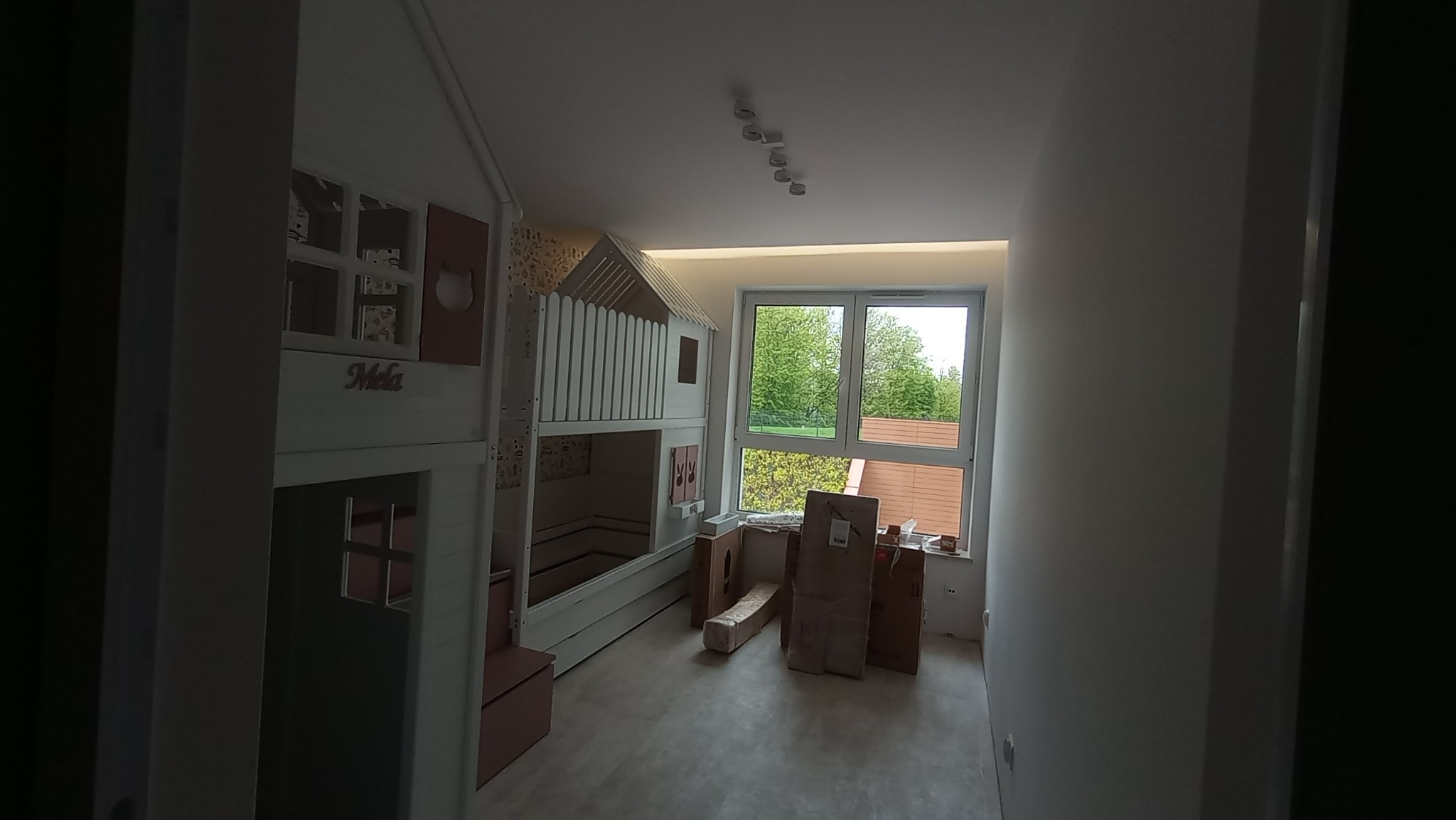 Pokój dziecięcy z zamontowanym łóżkiem piętrowym w kształcie domku, widok na okno z zielenią, w tle kartony i materiały budowlane, surowy stan wykończenia.