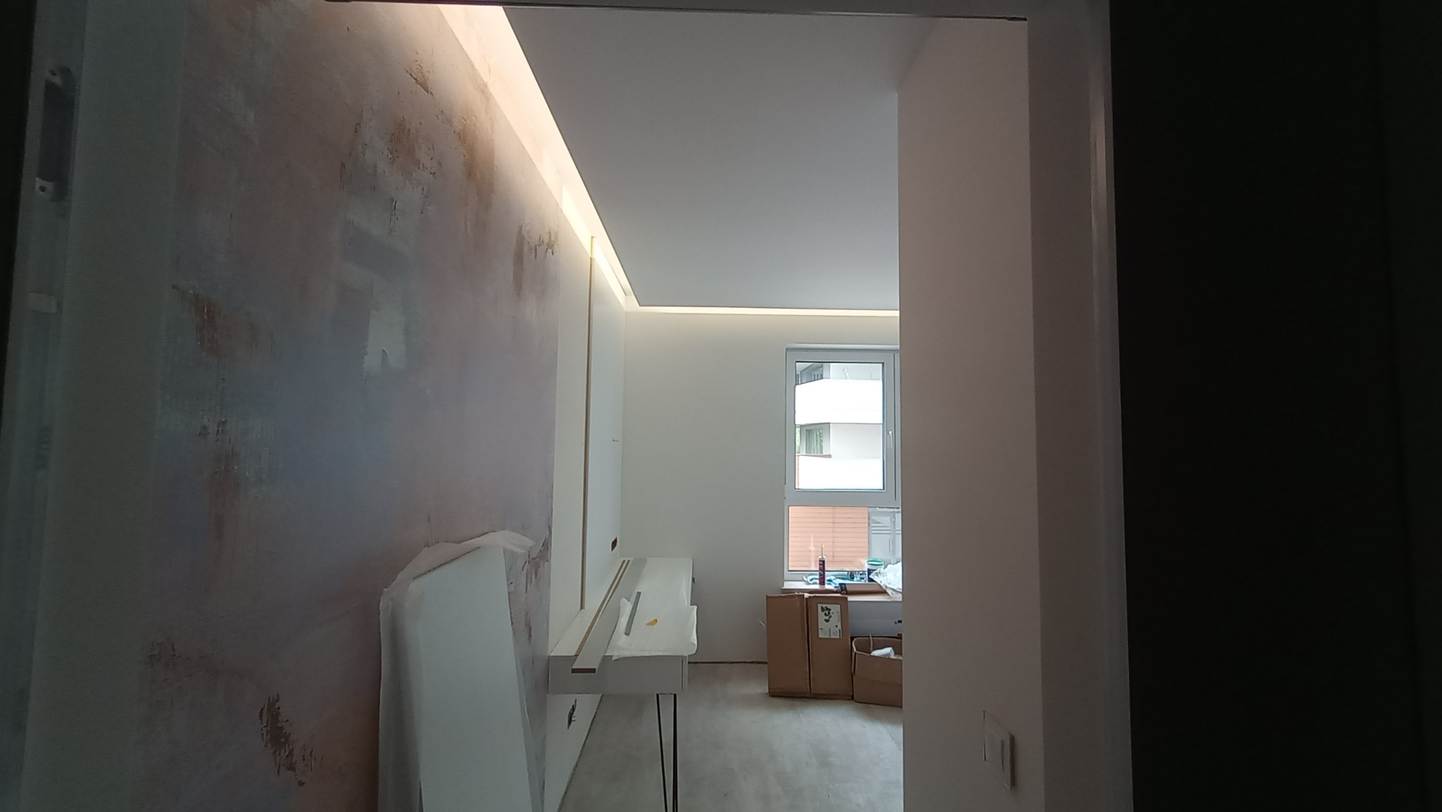 Pokój w trakcie remontu z dekoracyjną ścianą w odcieniach szarości i beżu, podświetlany sufit LED, widoczne kartony i elementy wyposażenia w tle, ujęcie z perspektywy wejścia.