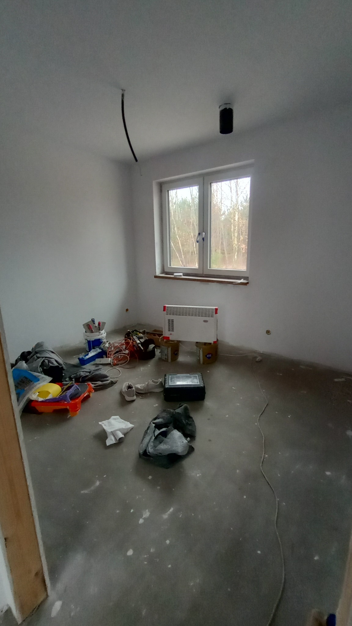 Pokój w trakcie prac wykończeniowych: świeżo pomalowane ściany na biało, surowa wylewka na podłodze z widocznymi plamami, rozstawione narzędzia i materiały malarskie, grzejnik elektryczny pod oknem.