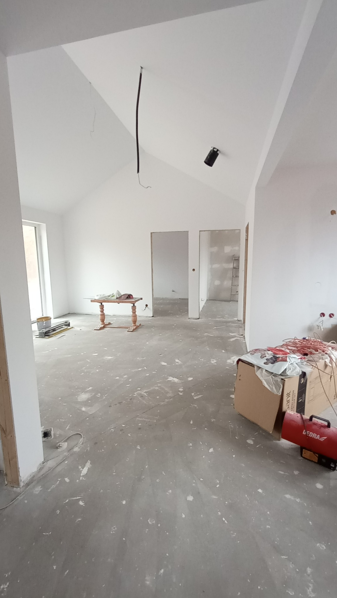 Wnętrze w trakcie remontu: surowe ściany pomalowane na biało, widoczne kable zwisające z sufitu, niezagruntowana podłoga z resztkami farby, grzejnik elektryczny i kartony w rogu pomieszczenia...