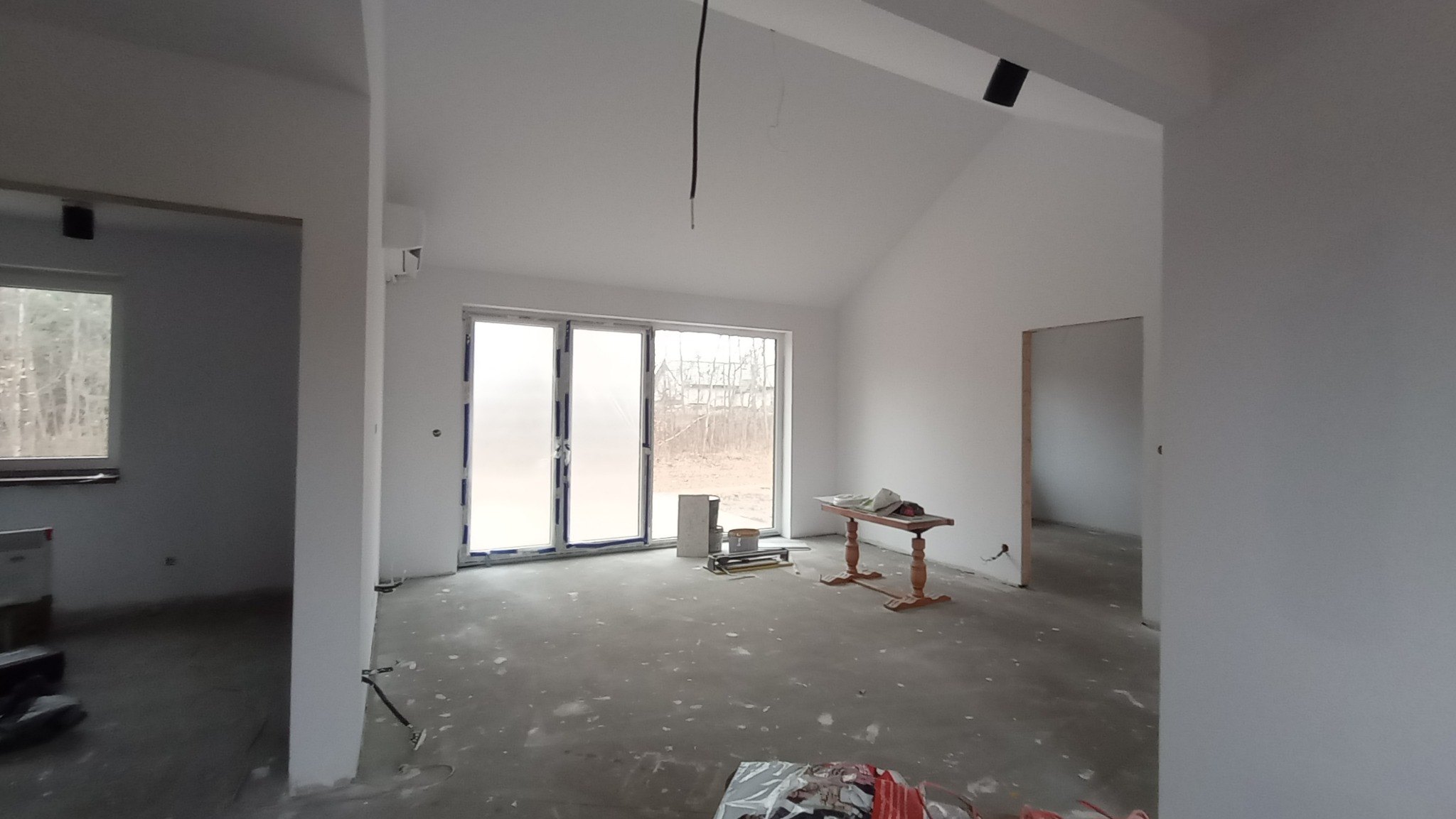 Wnętrze w stanie deweloperskim po pracach przygotowawczych, widoczne białe ściany, szpachlowana podłoga, okno balkonowe z folią ochronną, drewniany stolik i materiały budowlane.