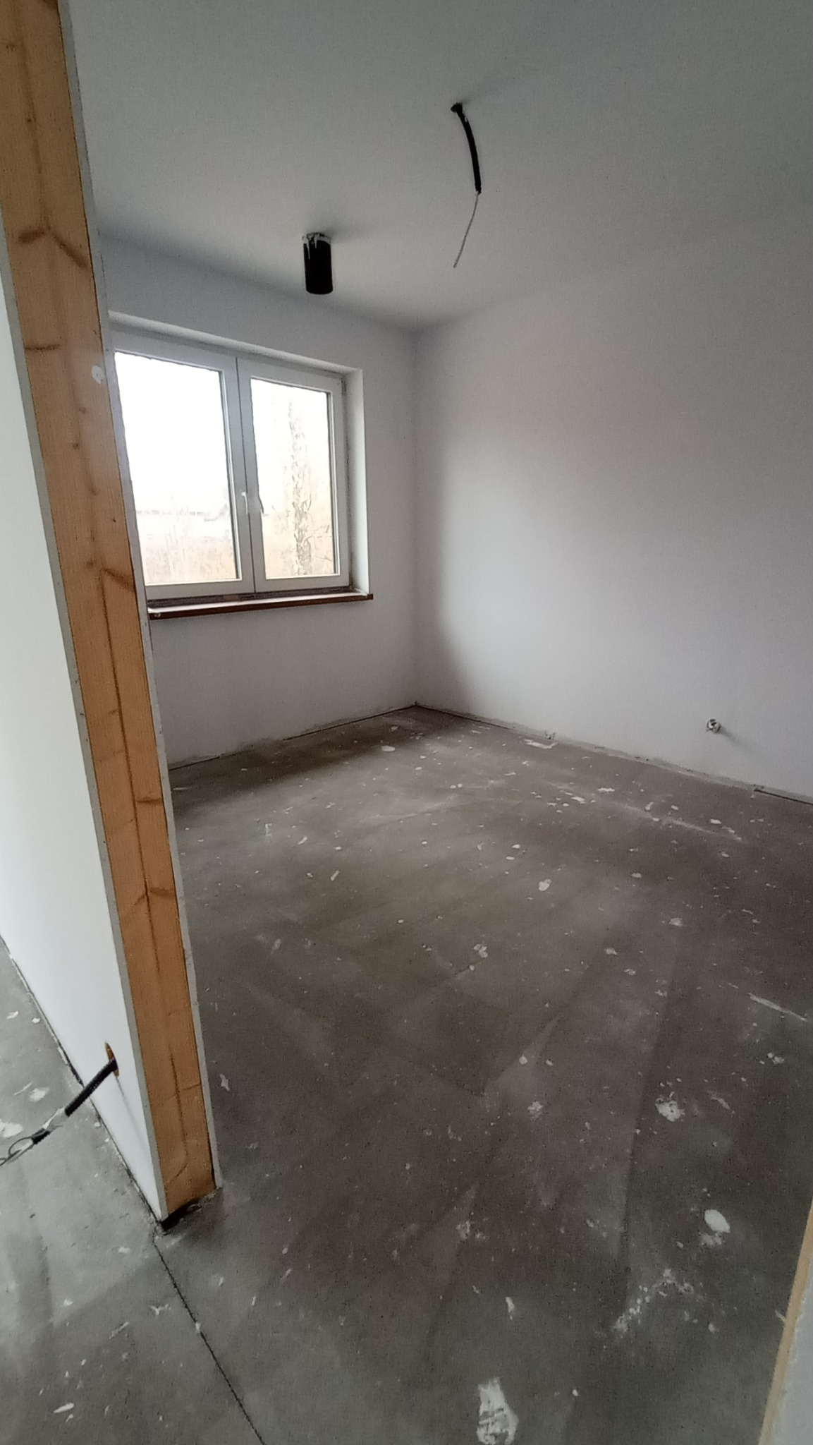 Wnętrze pokoju w trakcie remontu: surowe, białe ściany, szara wylewka na podłodze z widocznymi śladami po pracach, okno z drewnianą ramą i wystającymi kablami z sufitu.