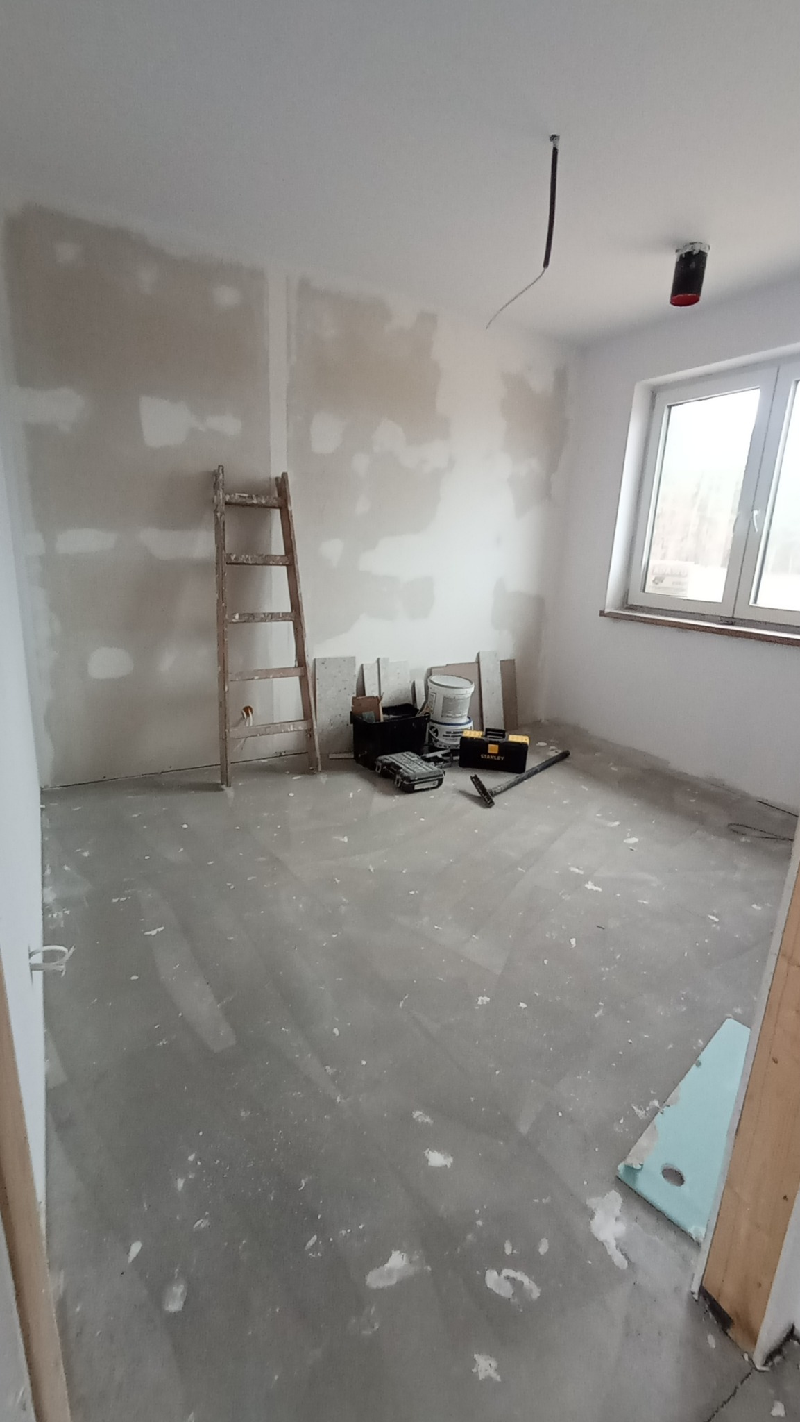 Pokój w trakcie remontu z widocznymi śladami po szpachlowaniu na ścianach, drewnianą drabiną opartą o ścianę, narzędziami i materiałami budowlanymi w tle oraz betonową wylewką na podłodze.