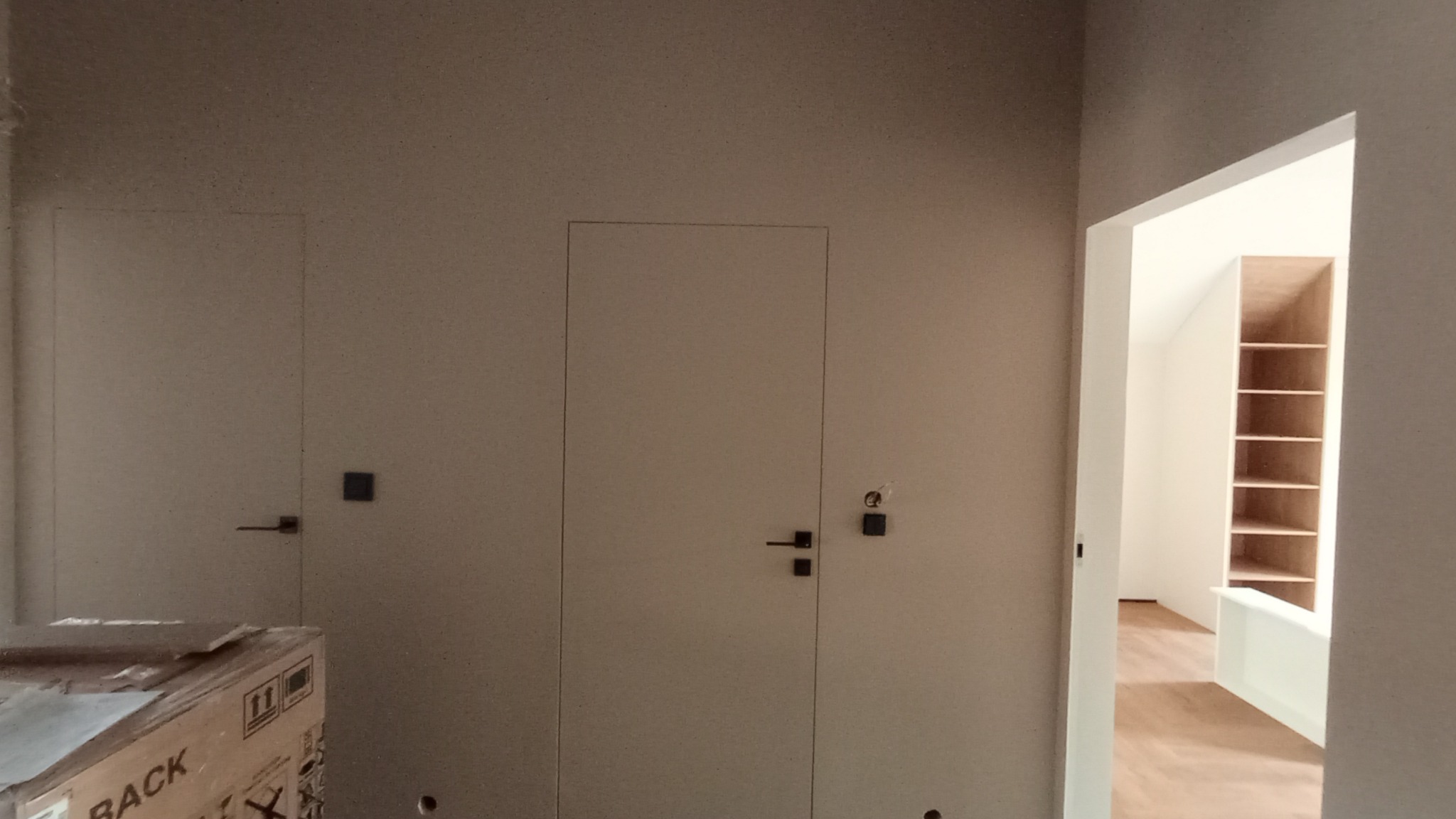 Wnętrze w trakcie wykańczania: ściany po szpachlowaniu, dwie ukryte drzwi z czarnymi klamkami, widok na wnętrze szafy wnękowej przez otwarte drzwi.