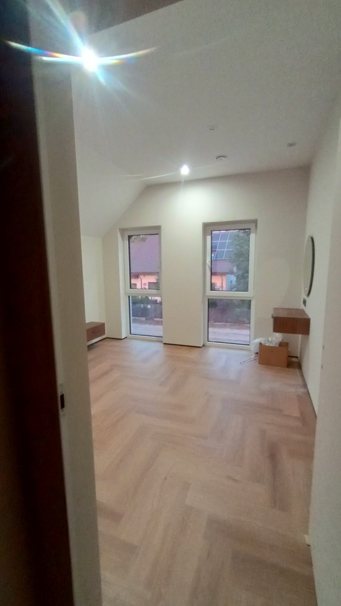 Pusty pokój po remoncie z parkietem ułożonym we wzór jodełki, świeżo pomalowanymi ścianami na biało i dwoma oknami z widokiem na domy z panelami słonecznymi.