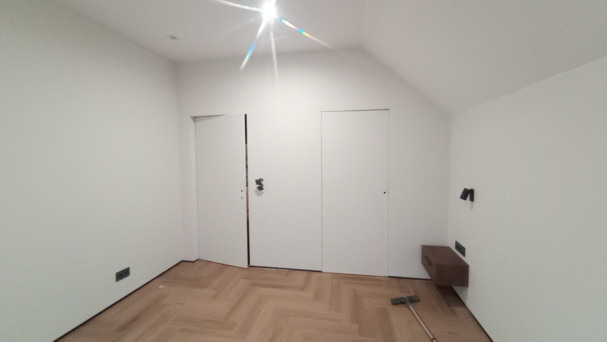 Białe wnętrze poddasza po remoncie z parkietem w jodełkę, dwojgiem białych drzwi, brązową szafką nocną i zamontowanymi kinkietami.