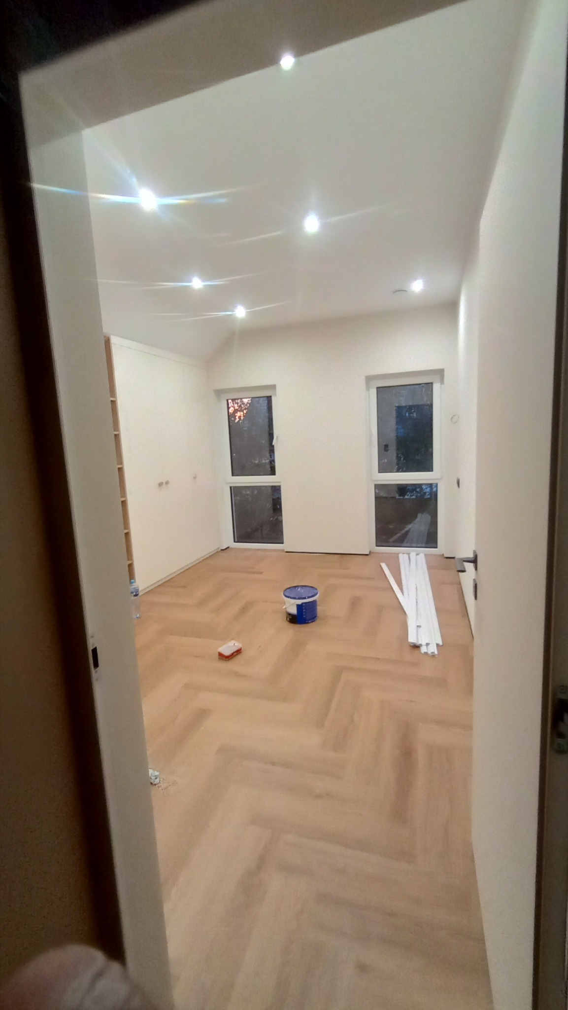 Wnętrze pokoju po remoncie, widoczne świeżo położone panele podłogowe w jodełkę, białe ściany, listwy przypodłogowe, okna z widokiem na zewnątrz, wiaderko z farbą i narzędzia malarskie. Oświetlenie...