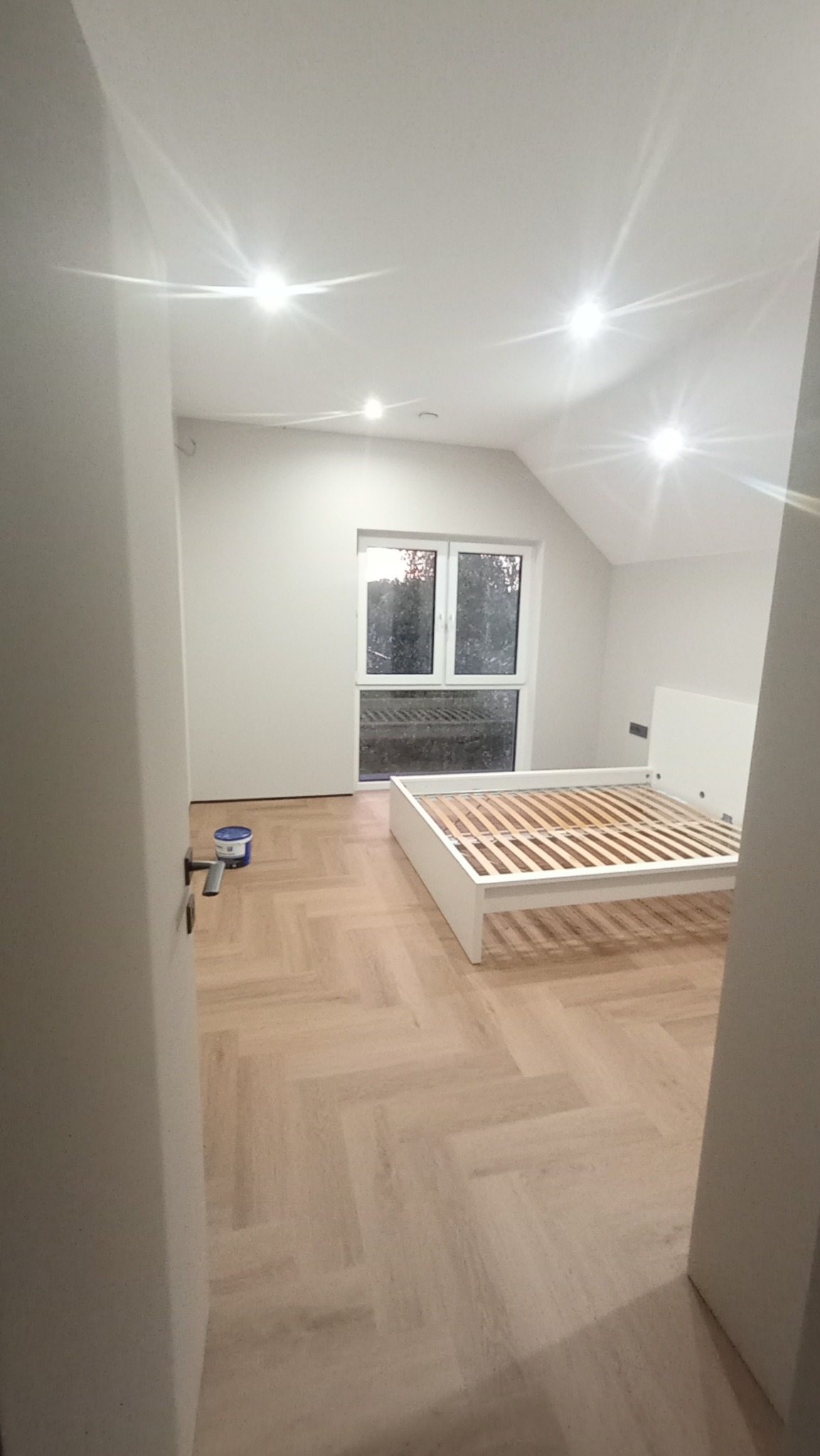 Jasne wnętrze pokoju z ułożoną podłogą w jodełkę, świeżo pomalowanymi ścianami i białym łóżkiem bez materaca; widoczne wiaderko z farbą przy drzwiach.