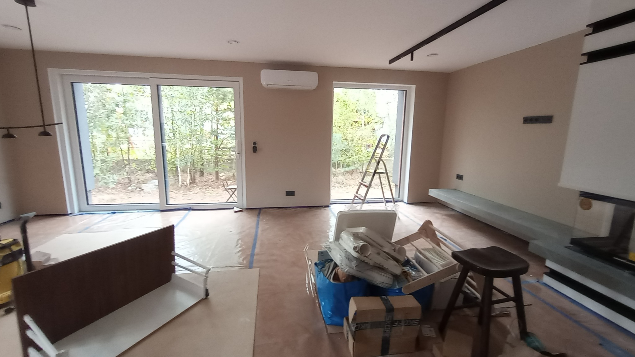 Wnętrze pokoju w trakcie prac wykończeniowych, zabezpieczona podłoga folią, widoczne narzędzia i materiały budowlane, drabina oparta o ścianę, ściany w kolorze beżowym, duże okna przesuwne...