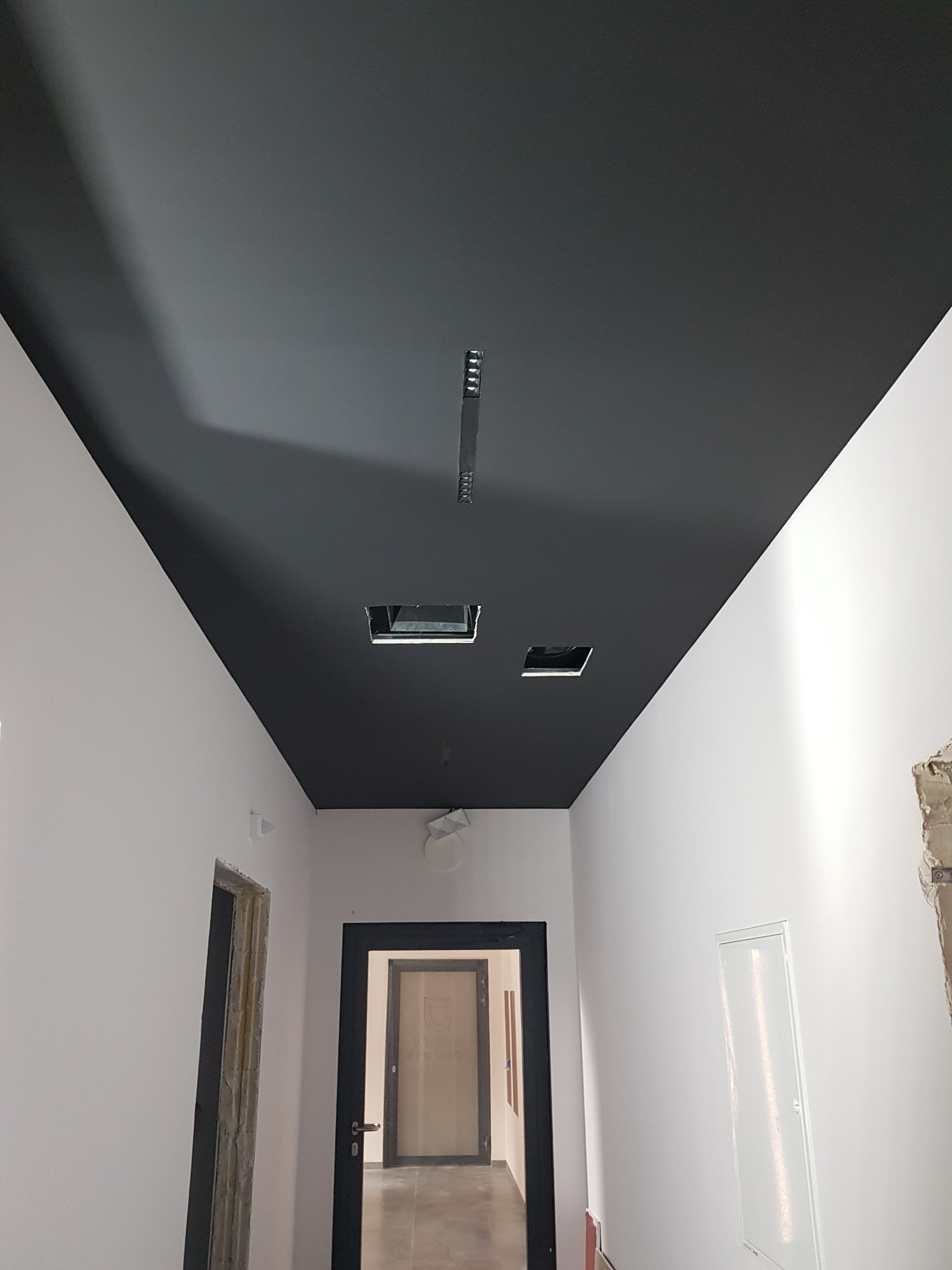 Korytarz w trakcie remontu z pomalowanym na czarno sufitem, widoczne otwory na oświetlenie i ściany przygotowane do dalszych prac wykończeniowych, surowe ościeżnice drzwiowe.