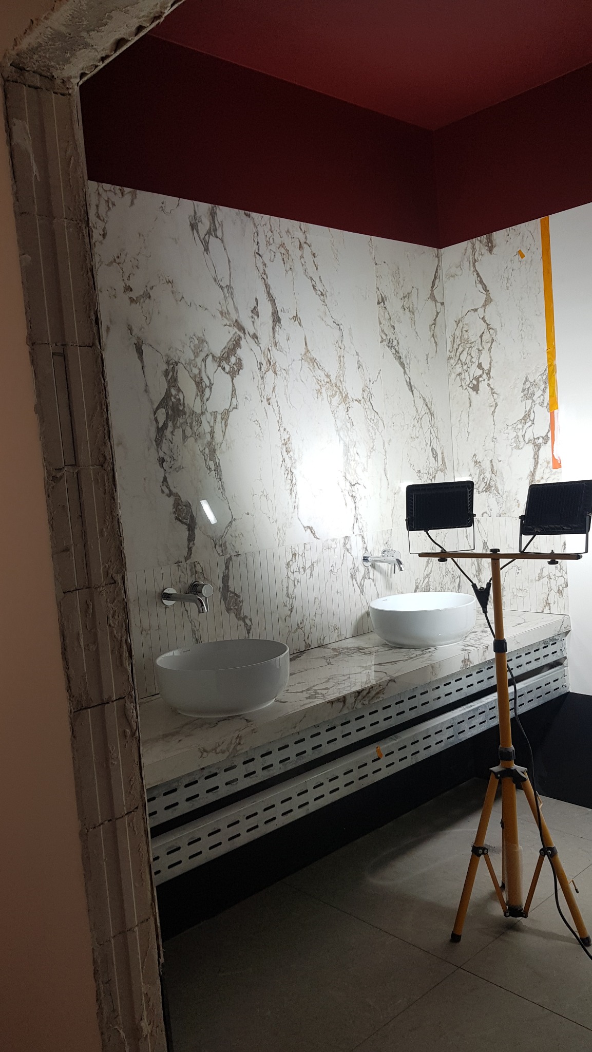 Łazienka w trakcie remontu: marmurowe płytki na ścianach, dwie białe umywalki, czerwony sufit, widoczne oświetlenie budowlane na statywie, surowy otwór drzwiowy.