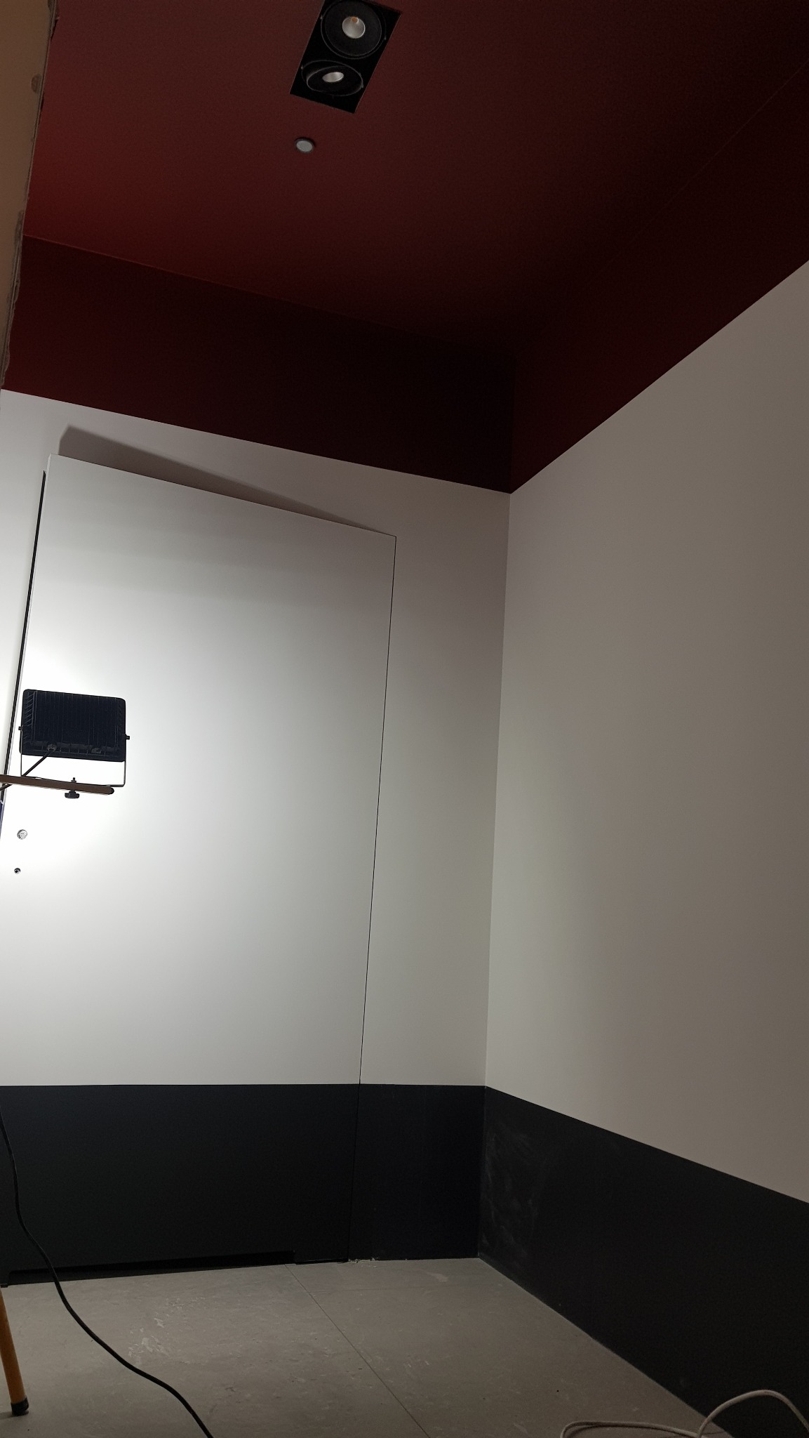 Wnętrze w trakcie prac wykończeniowych: białe ściany z czarnym pasem u dołu, oświetlone lampą budowlaną, z ciemnoczerwonym sufitem i widocznymi kablami na podłodze.