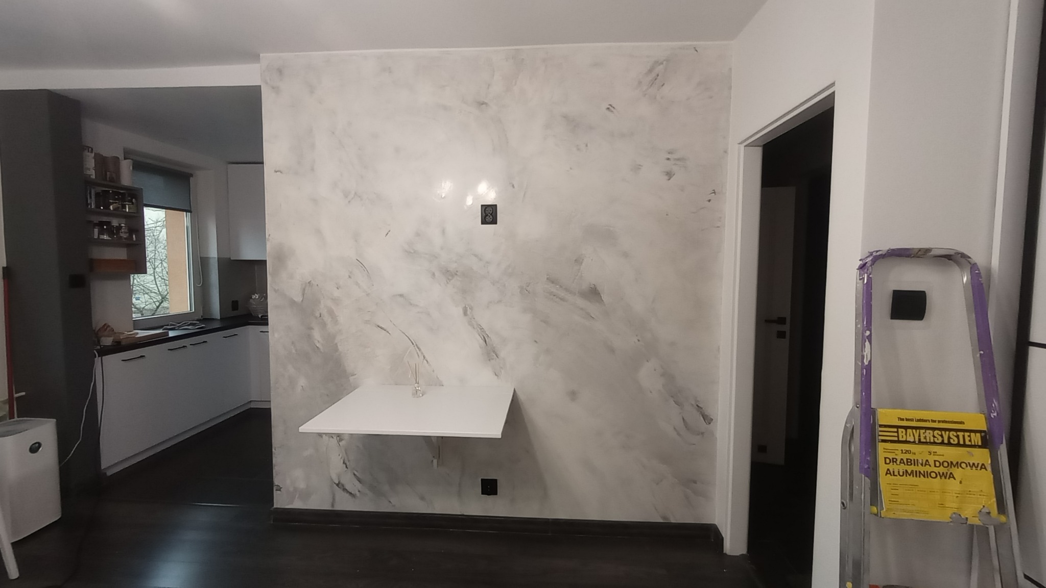 Ściana z dekoracyjną fakturą imitującą marmur w szaro-białych odcieniach, z zamontowanym składanym blatem, widoczna drabina aluminiowa i fragment kuchni w tle.