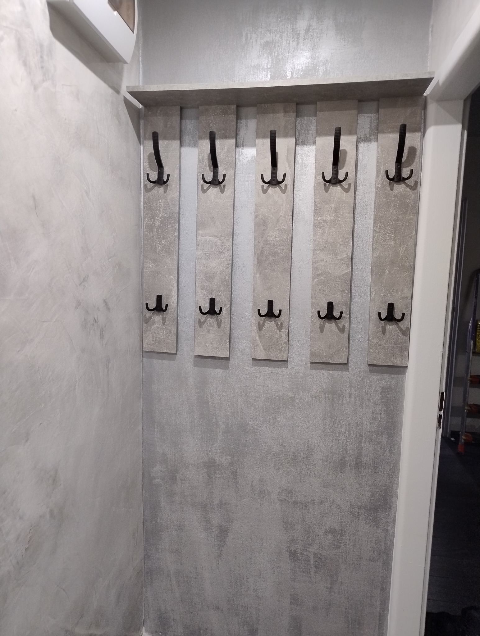 Ściana wykończona w szarym odcieniu z zamontowanym wieszakiem na ubrania z pięcioma podwójnymi, czarnymi haczykami i półką na górze.