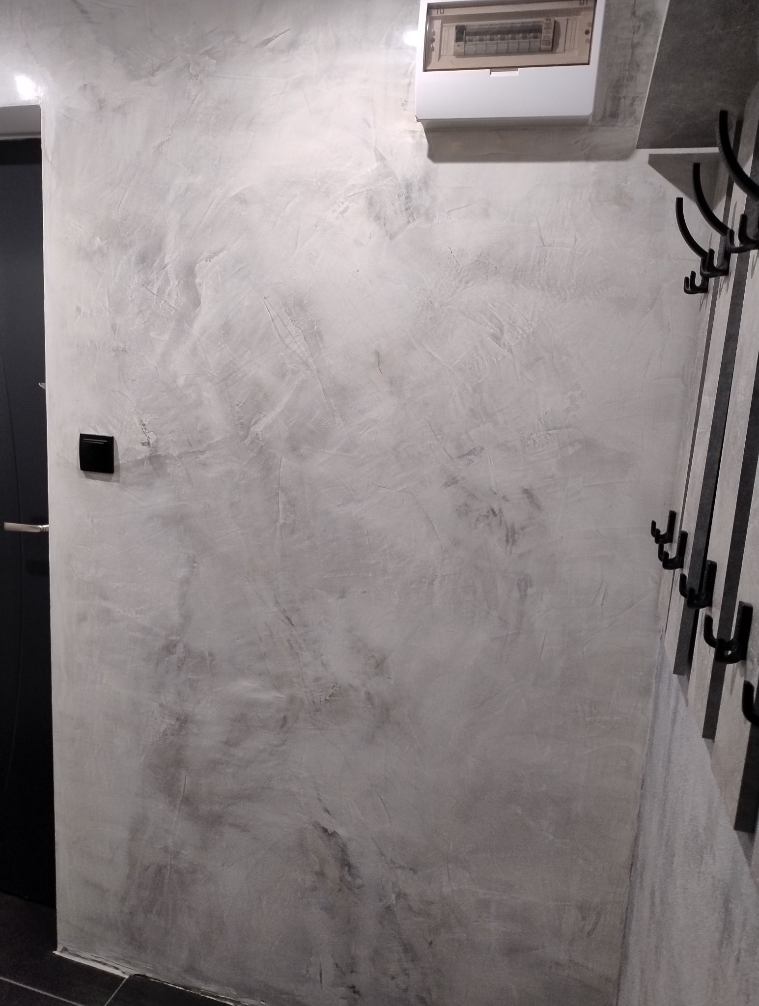 Ściana w korytarzu wykończona szarą, dekoracyjną gładzią imitującą beton, z widoczną skrzynką elektryczną i wieszakiem na ubrania.