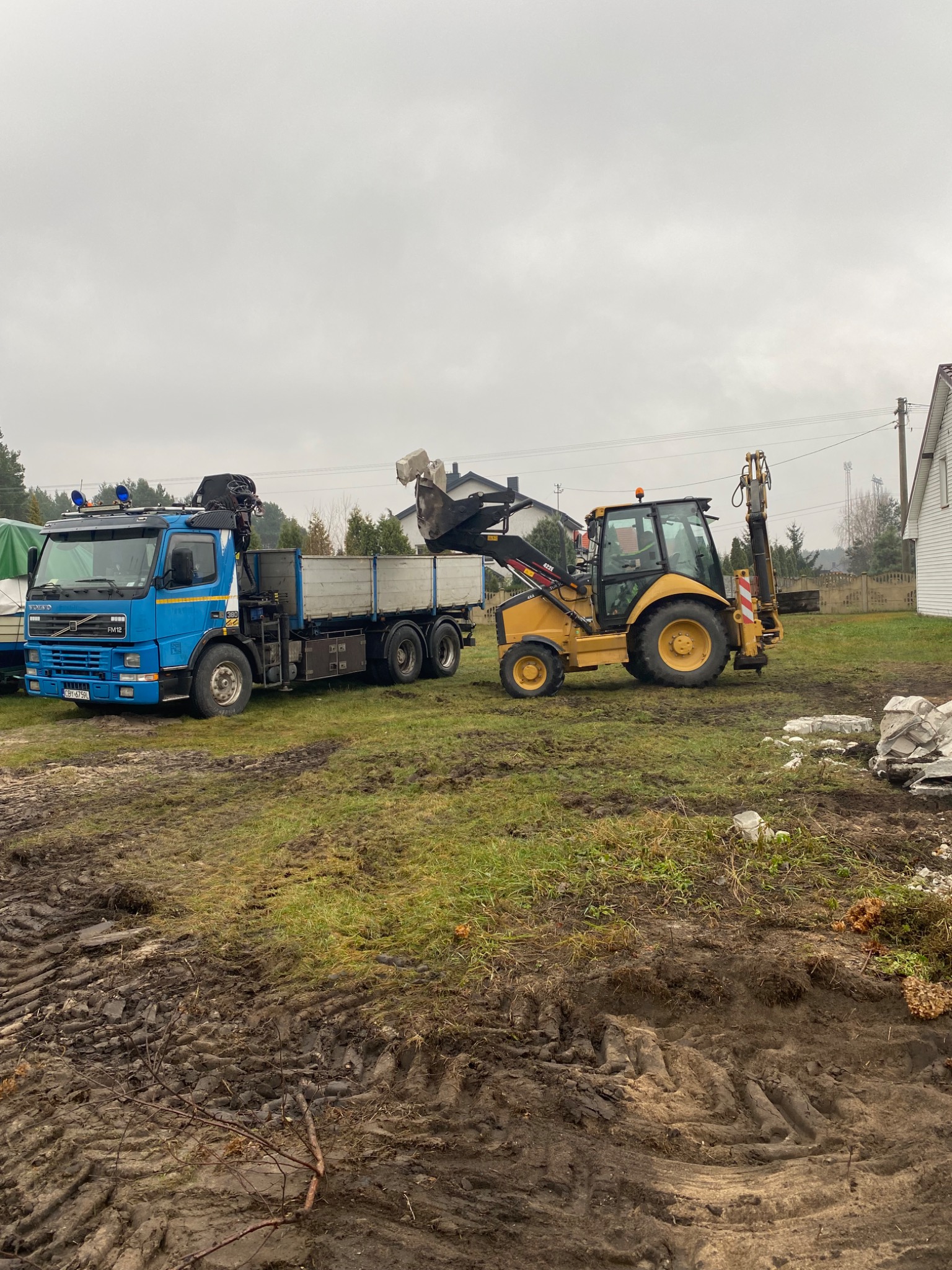 Niebieska ciężarówka Volvo z HDS i żółta koparko-ładowarka CAT na placu budowy podczas załadunku gruzu, widoczne ślady opon na błotnistym terenie, w tle dom jednorodzinny i pochmurne niebo.