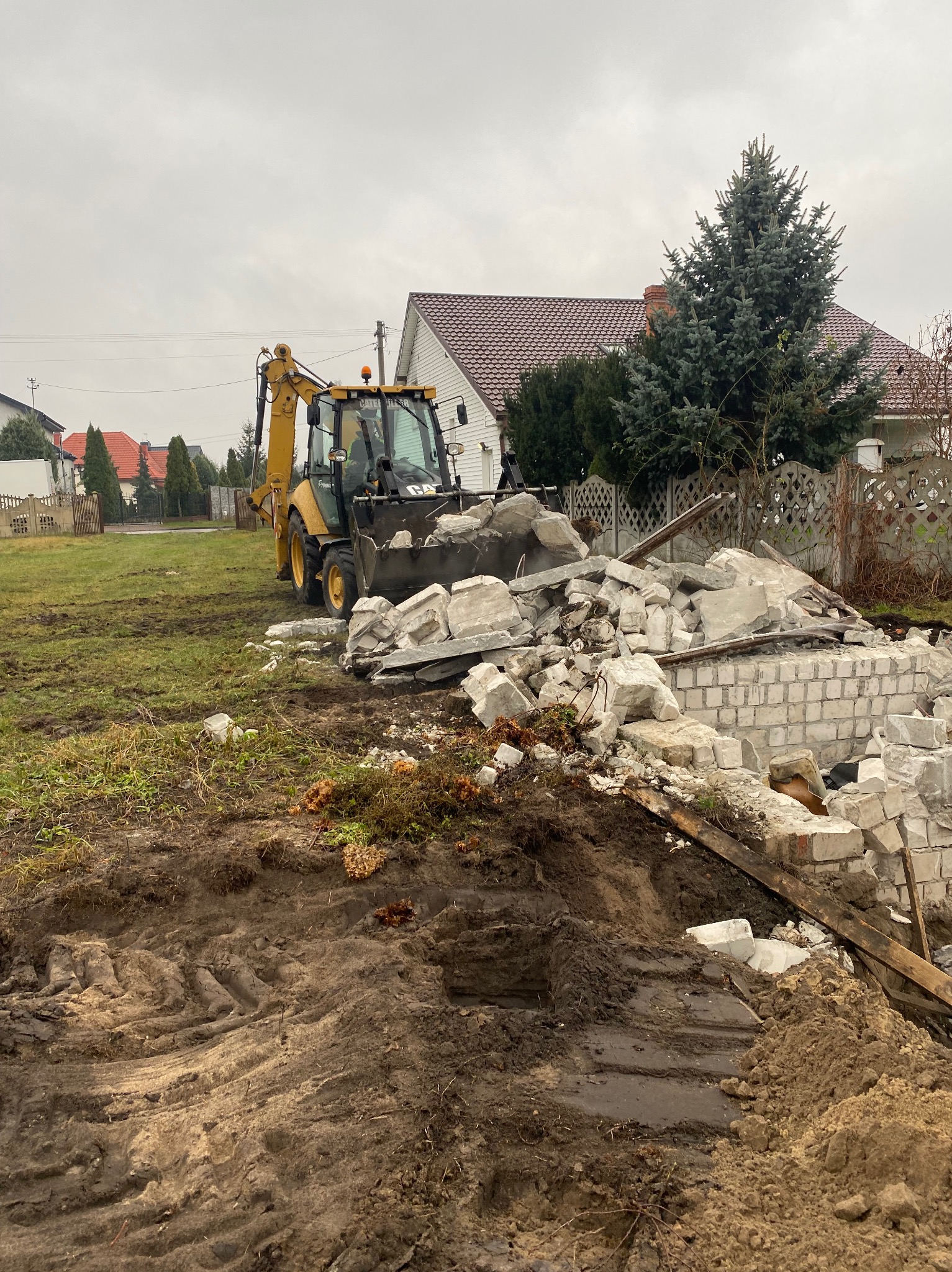 Żółta koparko-ładowarka CAT w trakcie rozbiórki murowanego budynku w miejscowości Białe Błota, z gruzem w łyżce i stertą cegieł w tle.