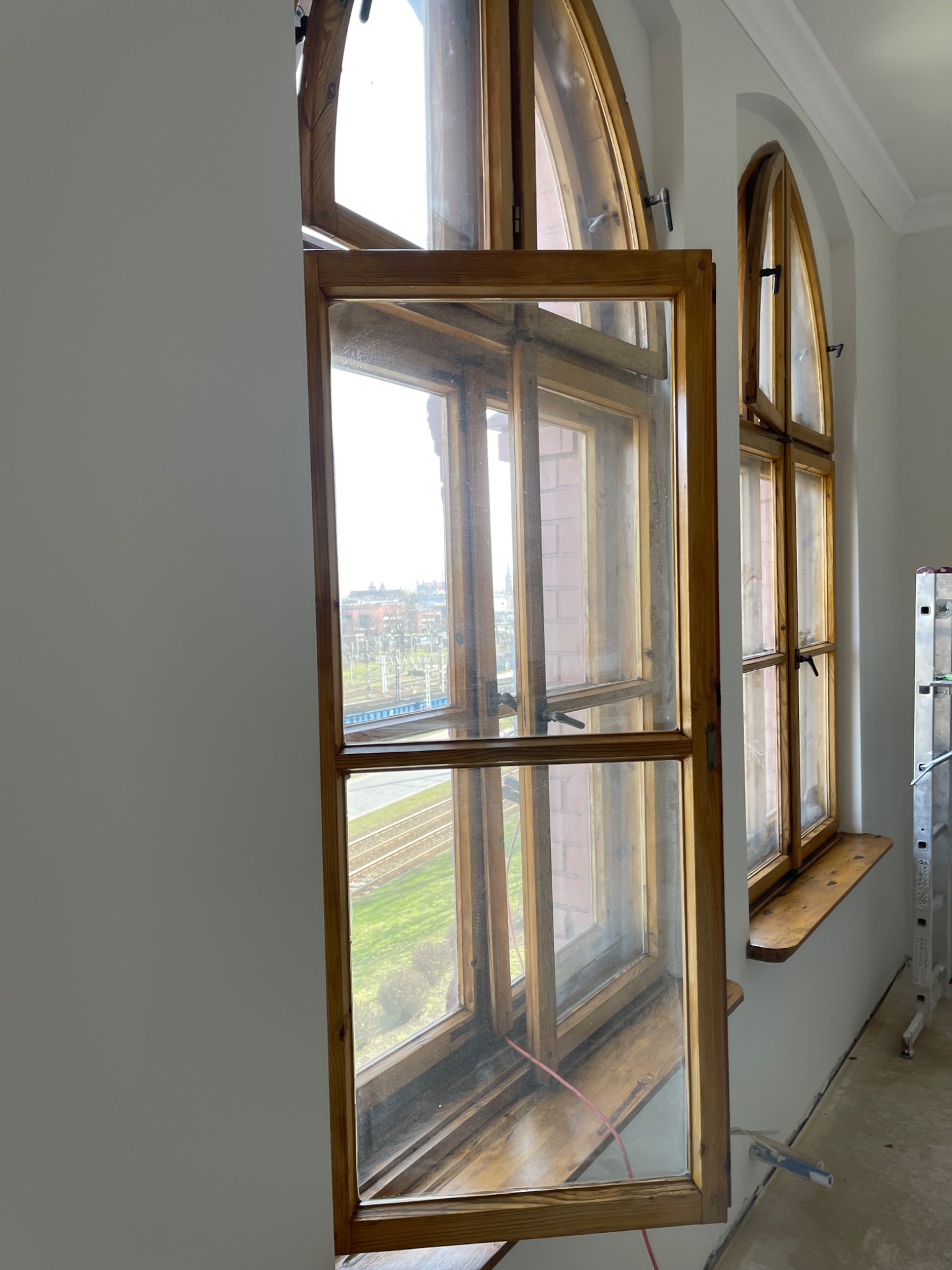 Otwarty drewniany okno z widokiem na tory kolejowe i budynki w oddali, obok drugie okno o łukowatym kształcie, drabina oparta o ścianę.