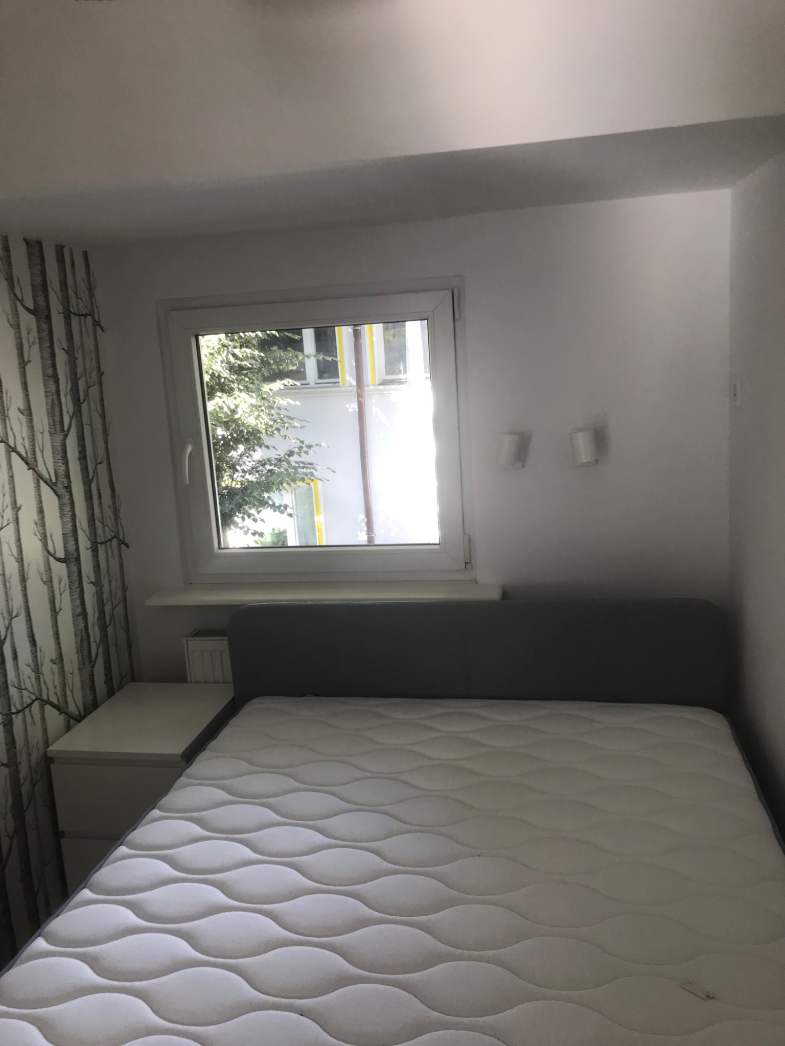 Pokój z łóżkiem, szafką nocną i oknem z widokiem na drzewa; widoczne dwa kinkiety na ścianie.