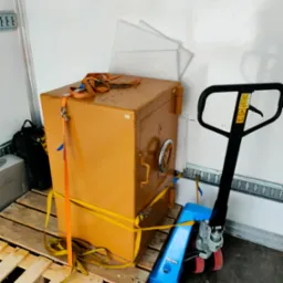 Sejf koloru musztardowego zabezpieczony pasami transportowymi na palecie, obok wózek paletowy, wewnątrz pojazdu transportowego.