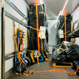 Kamado Kazimierz Zając - Wnętrze furgonetki z zabezpieczonymi meblami owiniętymi w czarną folię, spiętymi pomarańczowymi pasami transportowymi i wózkiem transportowym z niebieską ramą.