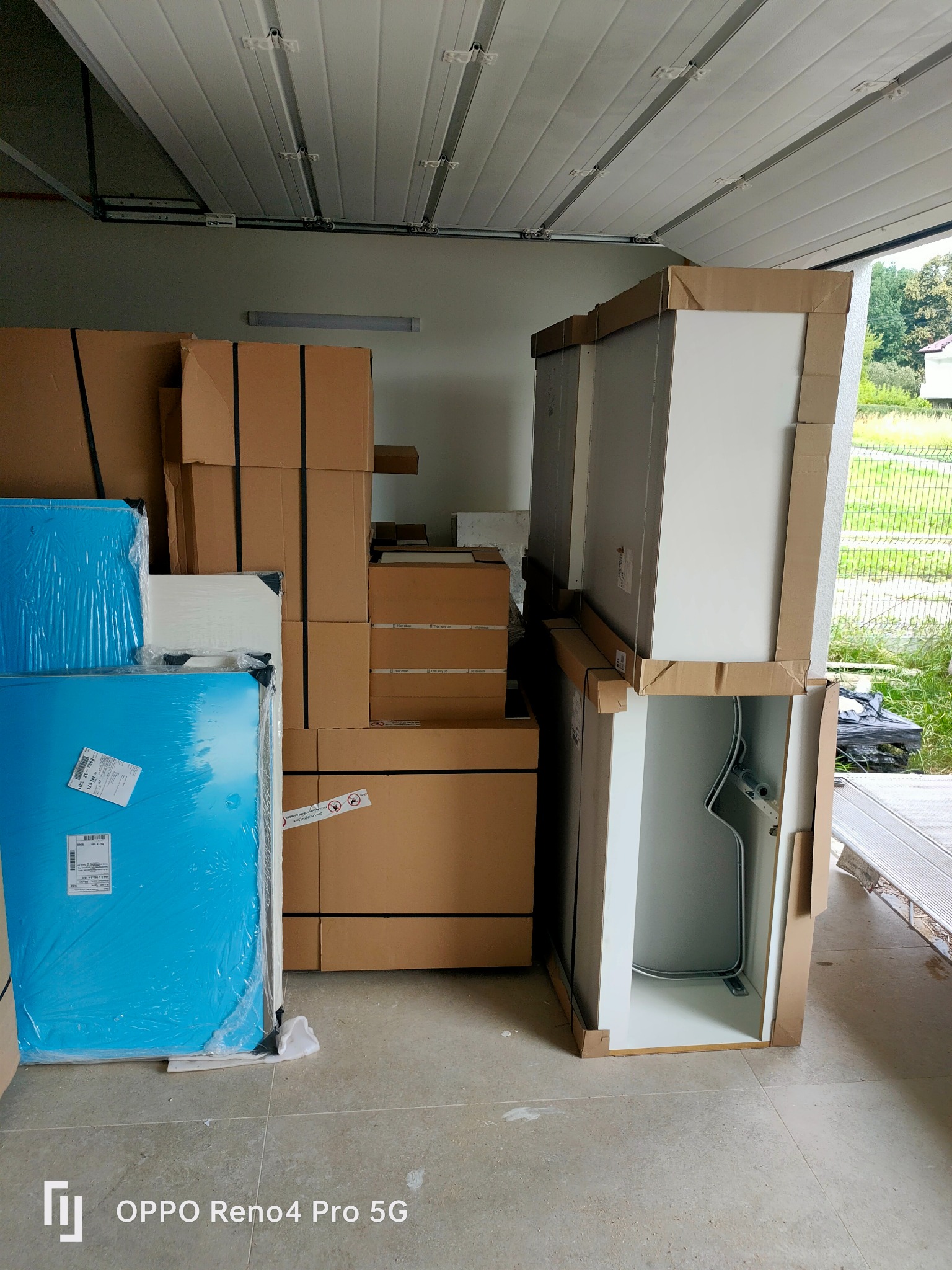 Paczki i meble łazienkowe w kartonach, przygotowane do transportu, w garażu z widocznym fragmentem posesji za oknem.