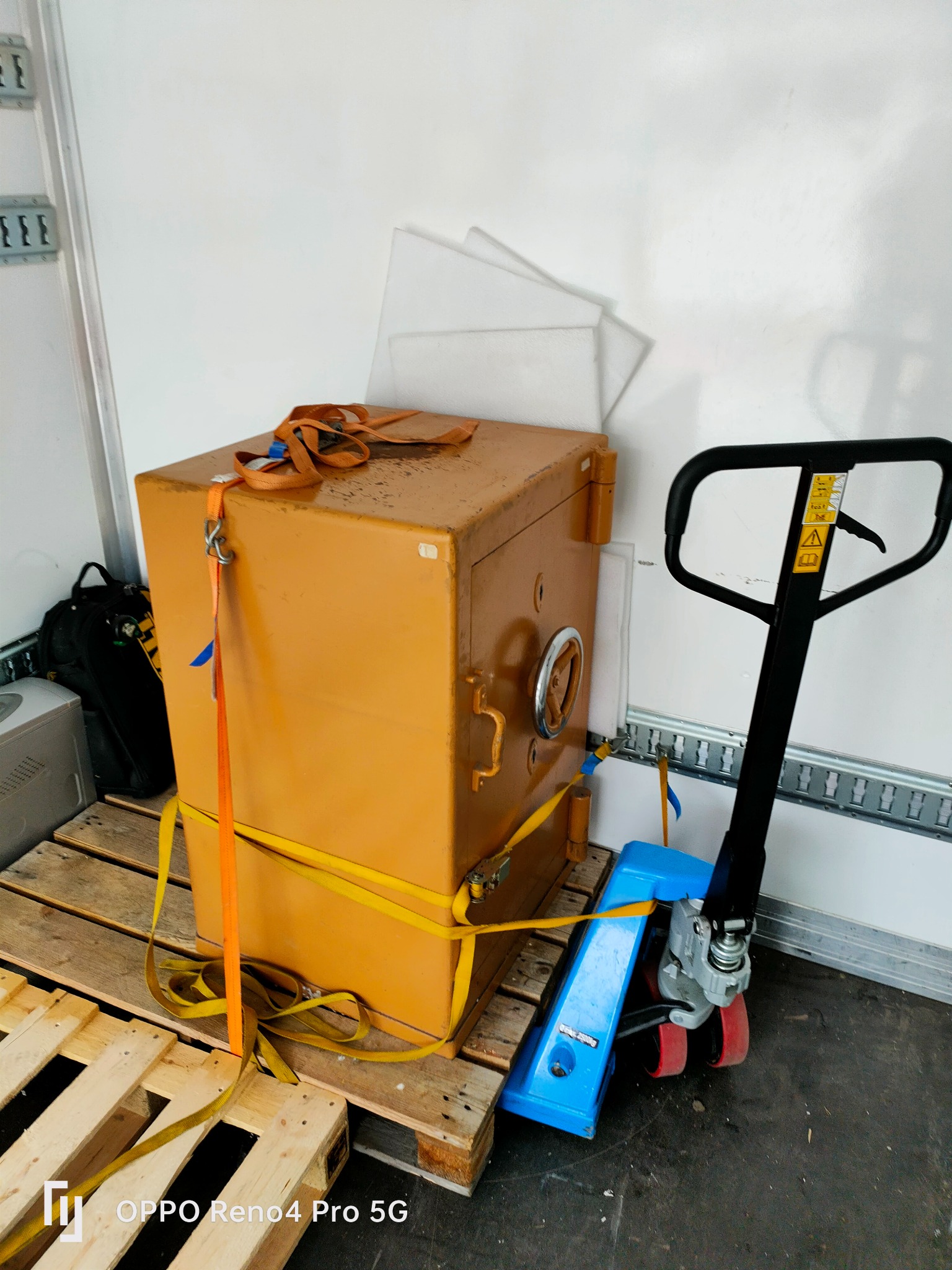 Sejf koloru musztardowego zabezpieczony pasami transportowymi na palecie, obok wózek paletowy, wewnątrz pojazdu transportowego.