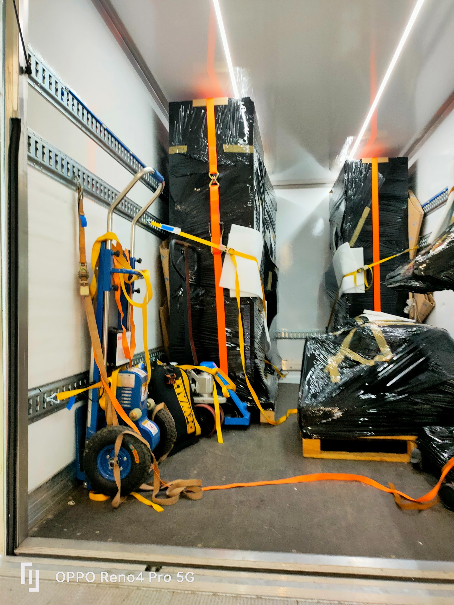 Wnętrze furgonetki z zabezpieczonymi meblami owiniętymi w czarną folię, spiętymi pomarańczowymi pasami transportowymi i wózkiem transportowym z niebieską ramą.