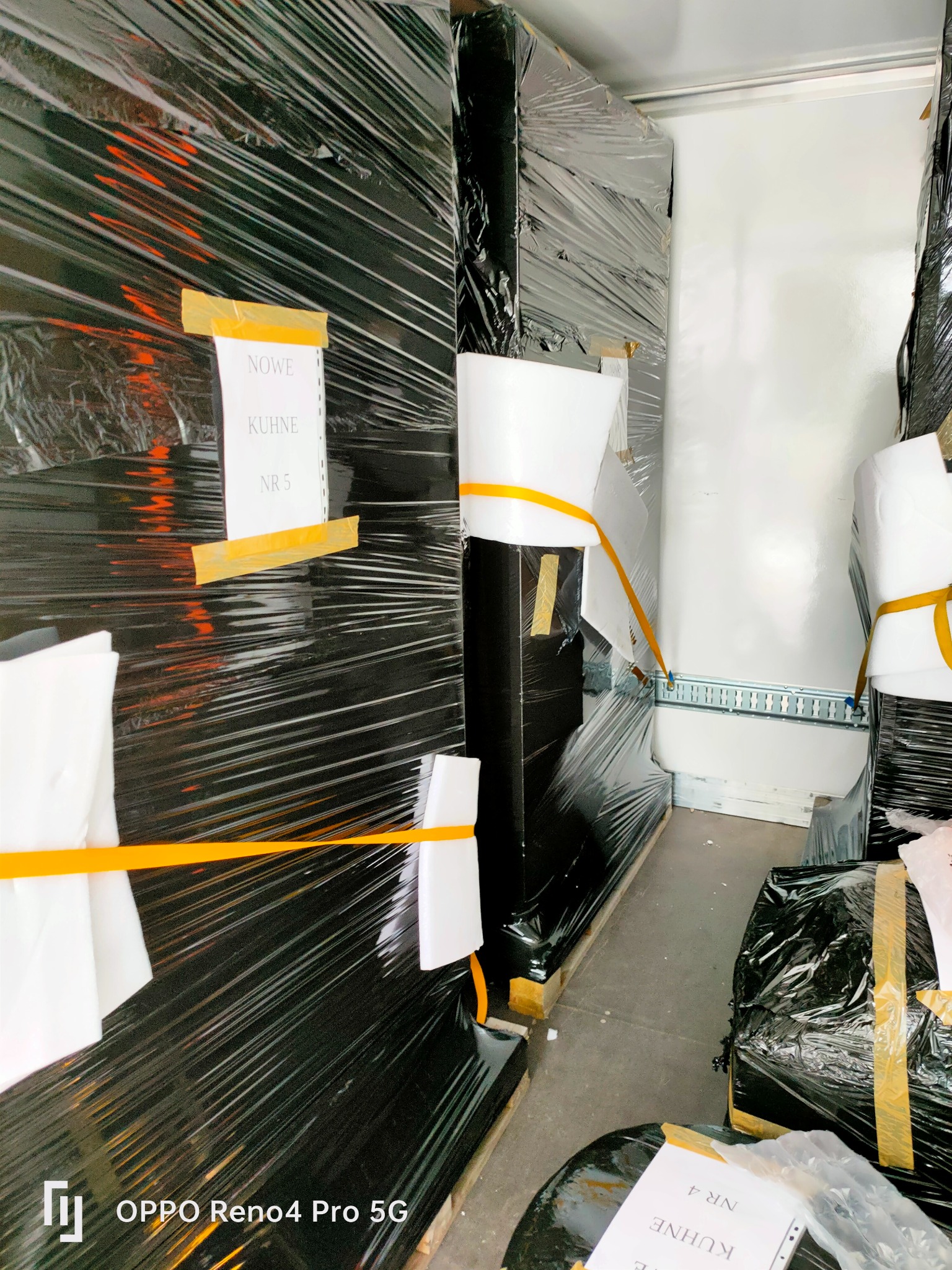 Wnętrze furgonetki wypełnionej meblami i sprzętem AGD, owiniętymi w czarną folię stretch i zabezpieczonymi taśmą oraz kartkami z napisem 'NOWE KUHNE NR 5' i 'KUHNE NR 4'.