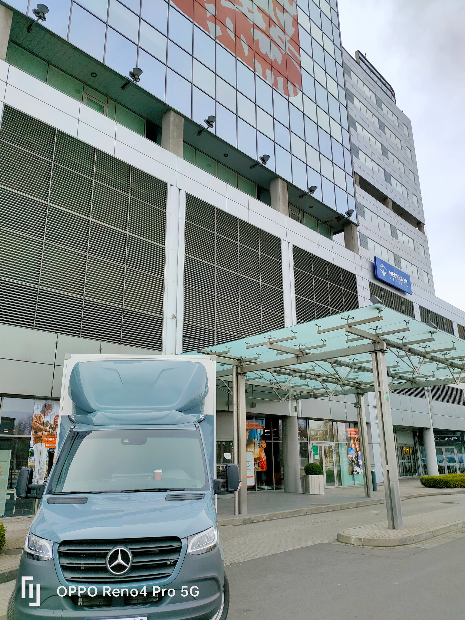 Błękitny bus dostawczy Mercedes Sprinter z nadbudową, zaparkowany przed nowoczesnym biurowcem Medicover Centrum z przeszklonym wejściem i metalową konstrukcją zadaszenia.