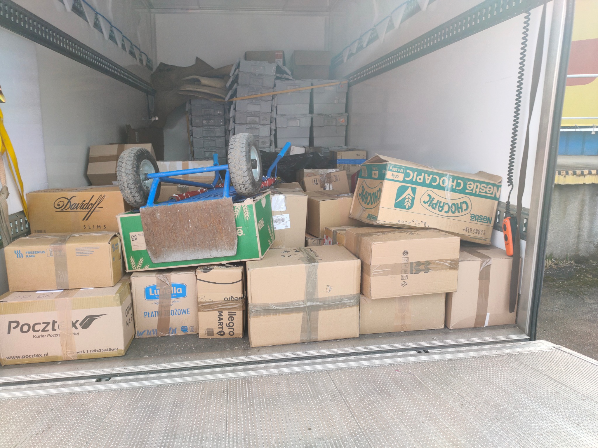 Wnętrze furgonetki załadowane kartonami różnych rozmiarów i z różnymi nadrukami, w tym logo Pocztex, Lubella, Davidoff i Nestle Chocapic, z widocznym wózkiem transportowym na górze, gotowe...