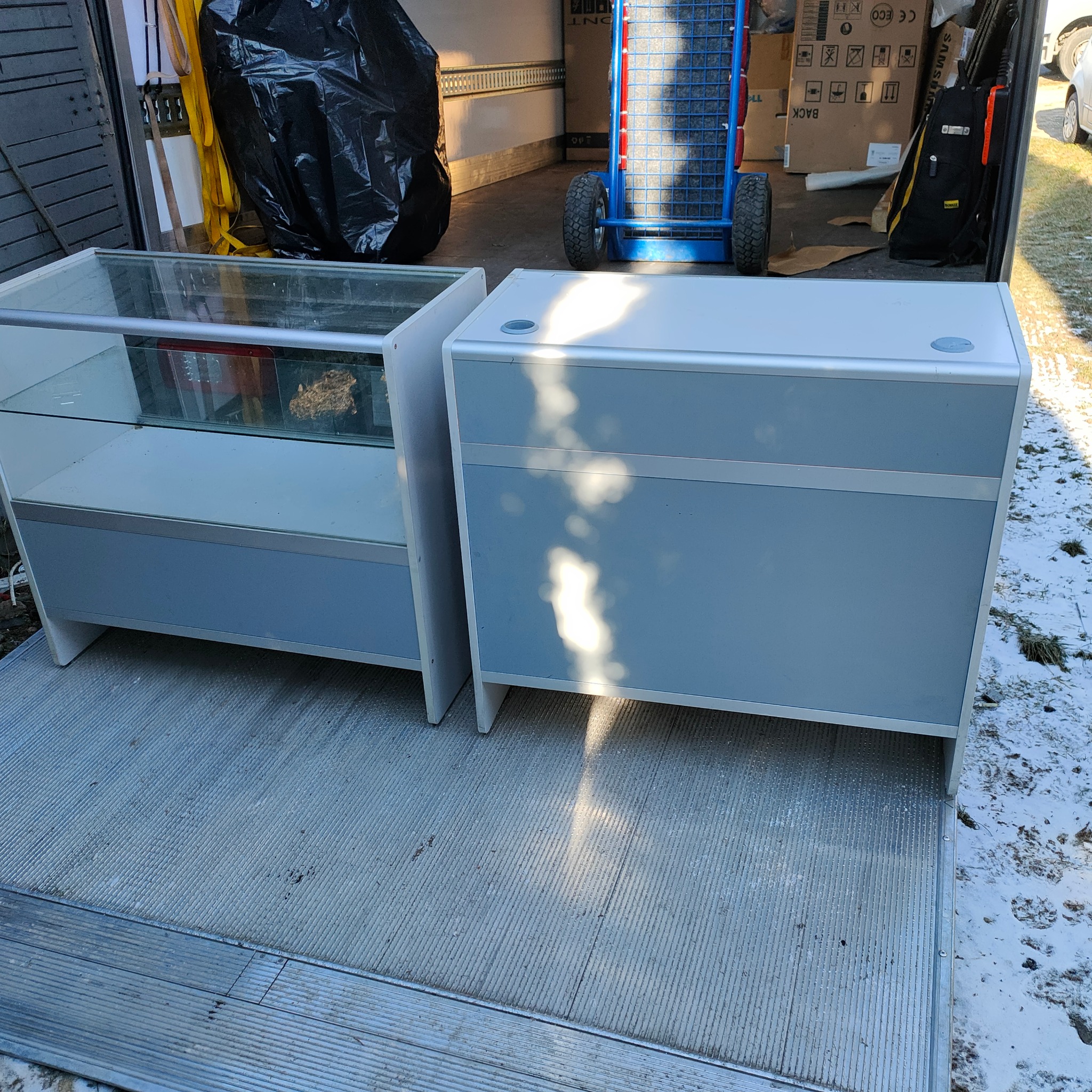 Dwa meble, witryna szklana i szafka, umieszczone na rampie busa, w tle wózek transportowy i zabezpieczony czarnym workiem ładunek.