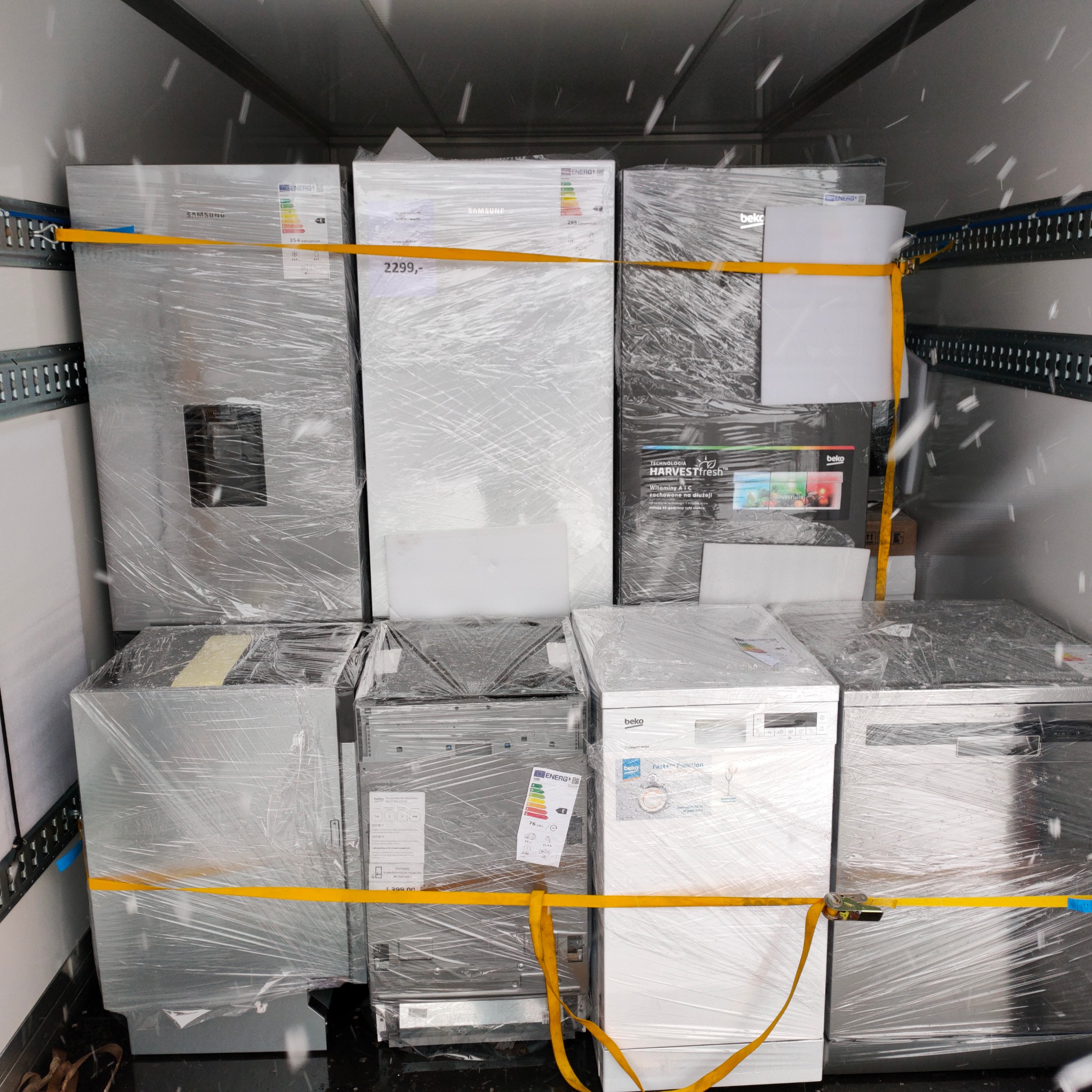 Wnętrze busa wypełnione sprzętem AGD (lodówki, zmywarki, pralki) owiniętym folią i zabezpieczonym żółtymi pasami transportowymi, w tle metalowe ściany pojazdu.