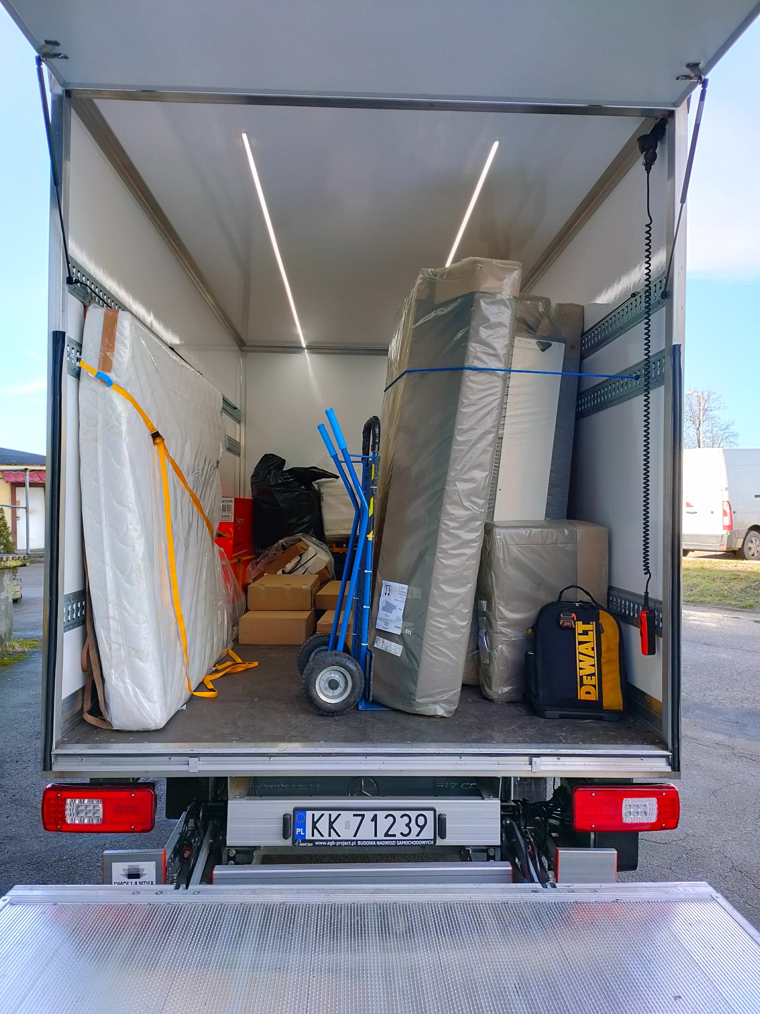 Wnętrze busa transportowego, załadowane meblami i kartonami zabezpieczonymi folią i pasami, widoczny wózek transportowy i torba narzędziowa DeWalt, oświetlenie LED w suficie, rampa załadunkowa
