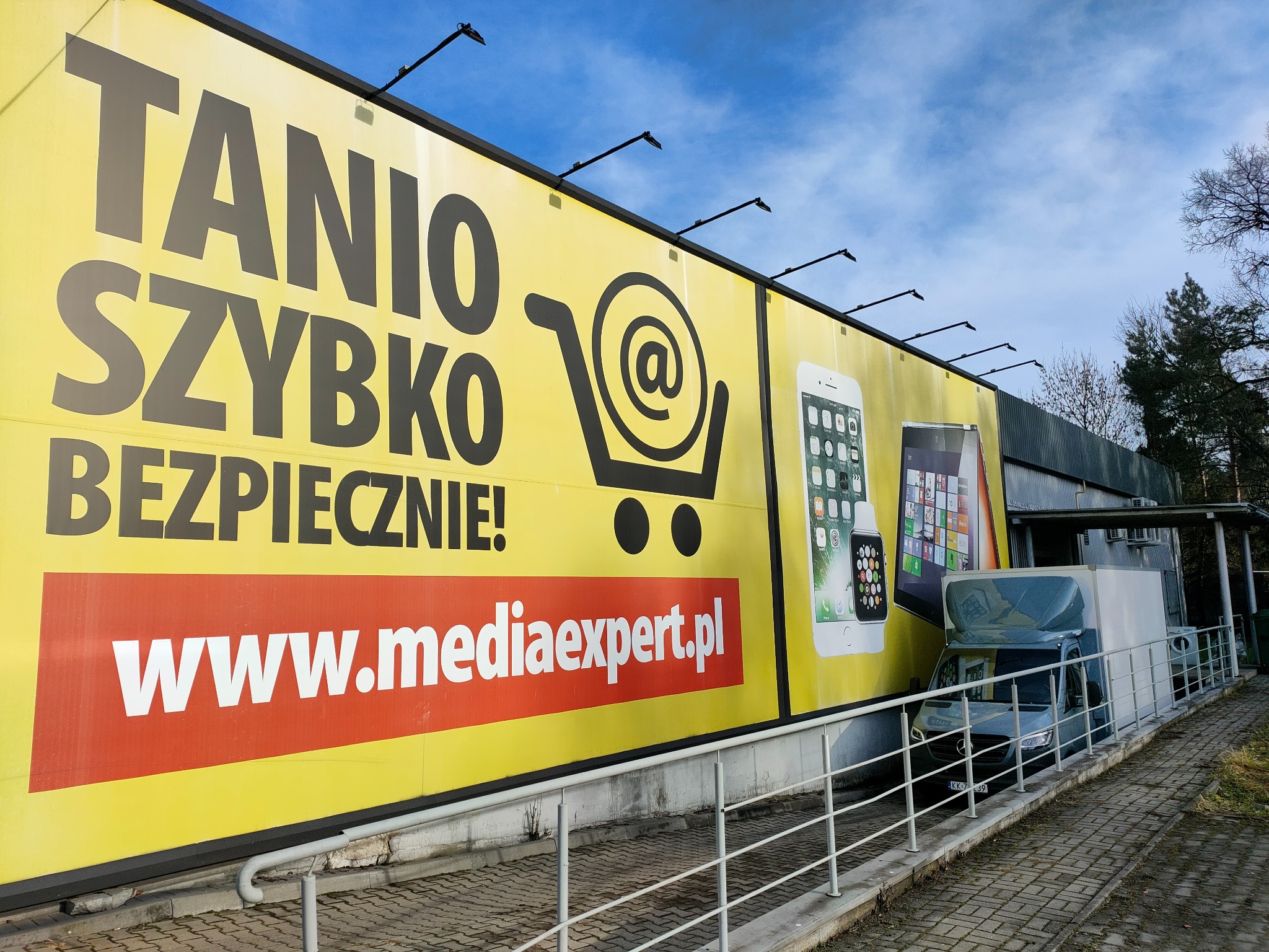 Reklama wielkoformatowa sklepu Media Expert z hasłami 'Tanio, Szybko, Bezpiecznie!' i adresem strony internetowej, obok zaparkowany bus dostawczy.