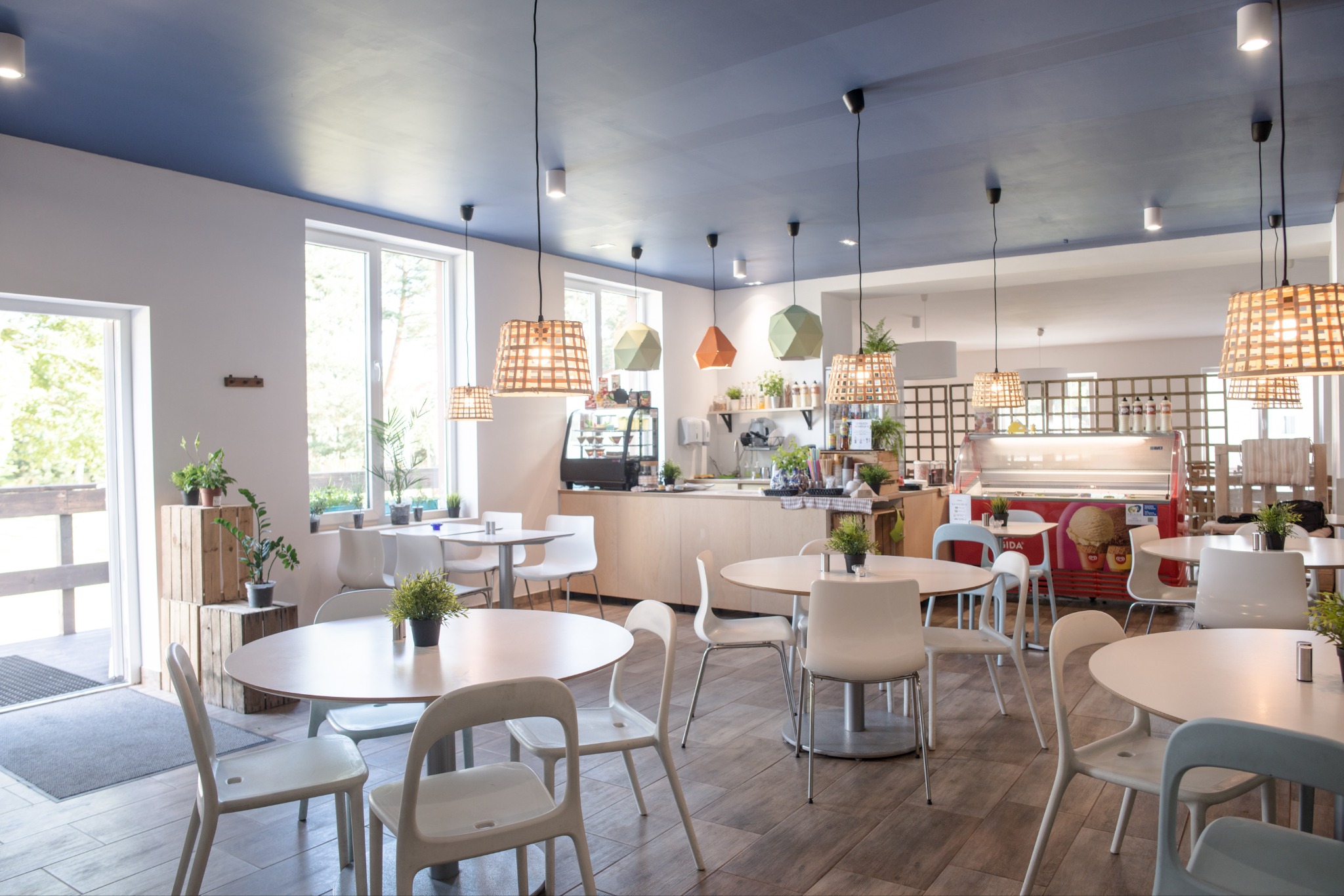 Jasne wnętrze stołówki ośrodka wypoczynkowego z okrągłymi stolikami, białymi krzesłami i dekoracyjnymi lampami wiszącymi o różnych kształtach, widok na zewnątrz przez otwarte drzwi.