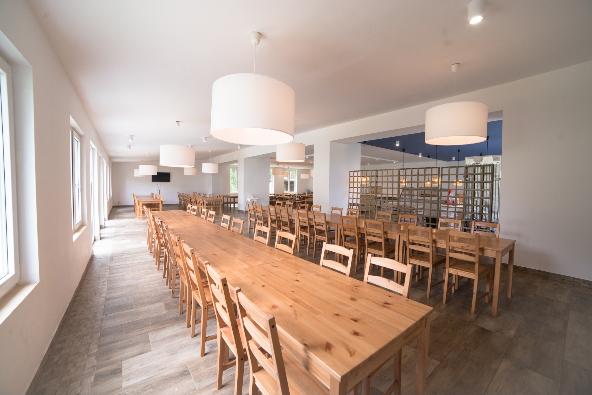 Jasna sala jadalna z długimi drewnianymi stołami i krzesłami, oświetlona okrągłymi lampami wiszącymi; widok na dalszą część sali przez ażurową ściankę działową.
