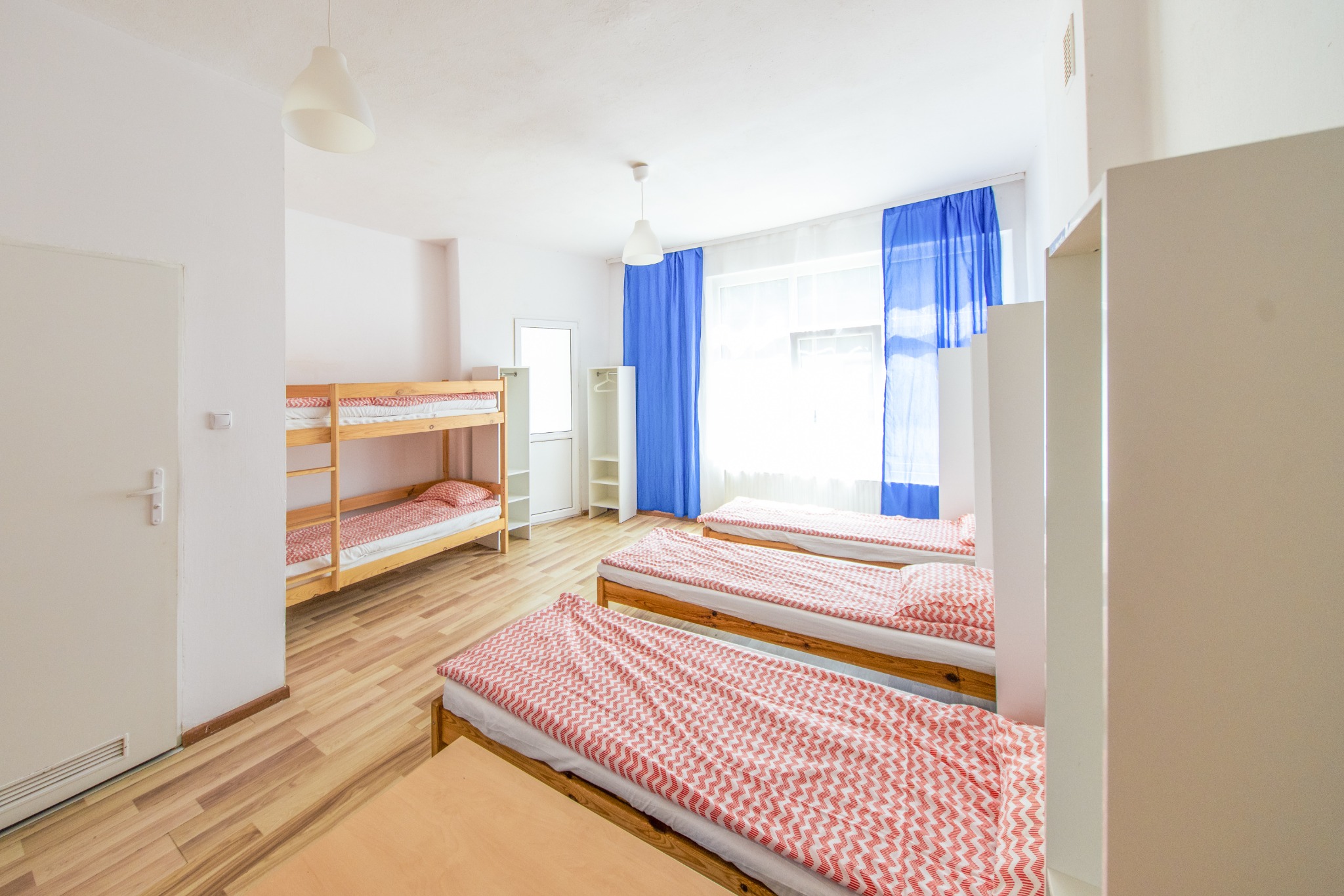 Wnętrze pokoju z łóżkami piętrowymi i pojedynczymi z pościelą w czerwono-biały zygzak, niebieskie zasłony w oknie, białe ściany i drzwi, drewniana podłoga.