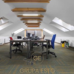 Grupa Esto Prawnicy i Pośrednicy - Przestronne biuro na poddaszu z oknami dachowymi, wyposażone w biurka, krzesła (jedno niebieskie), tablicę suchościeralną i grzejniki, z widocznym logo 'Grupa Esto' na pierwszym planie.