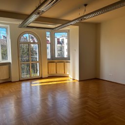 Grupa Esto Prawnicy i Pośrednicy - Puste, jasne pomieszczenie z parkietem w jodełkę, trzema oknami z widokiem na budynki i drzewa, oraz nowoczesnym oświetleniem sufitowym.