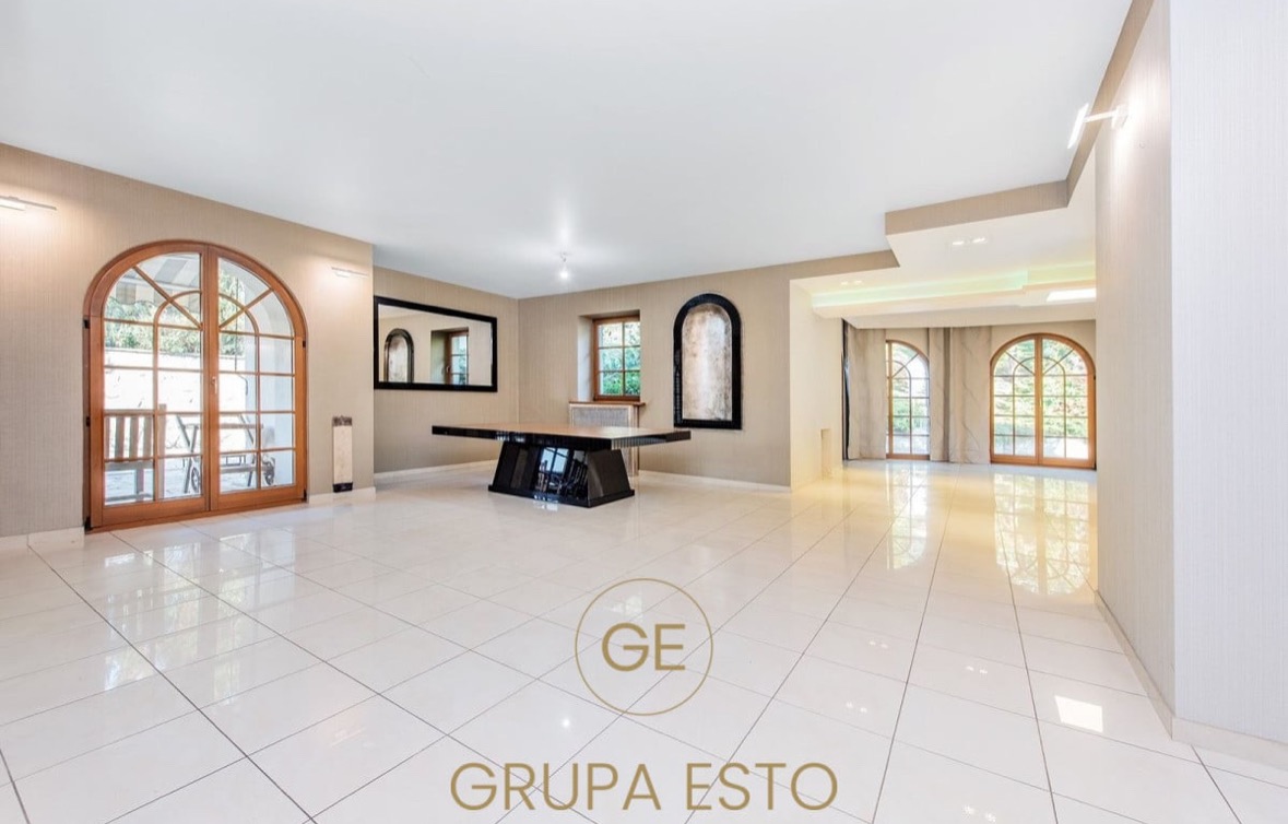 Przestronne, jasne wnętrze z dużymi, łukowymi oknami, błyszczącą podłogą z płytek i nowoczesnym stołem na środku, z logo firmy w dolnej części kadru.