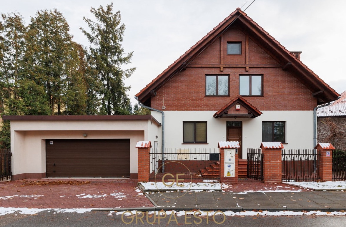 Dwukondygnacyjny dom z ceglaną elewacją na górze i białym tynkiem na dole, z garażem po lewej stronie, otoczony ogrodzeniem z ceglanymi słupkami i metalowymi przęsłami, częściowo pokryty śniegiem.